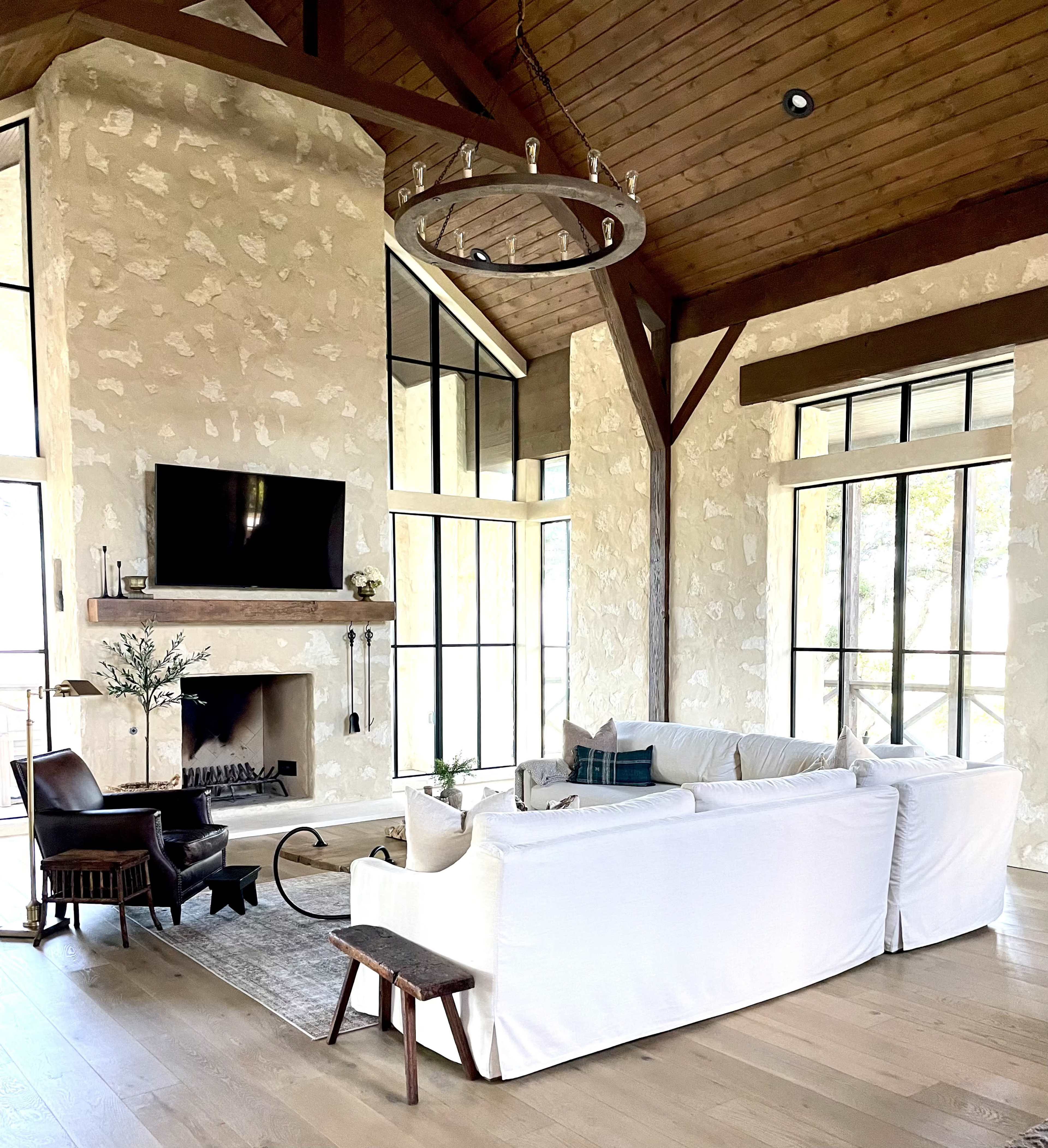 The image shows a modern living room featuring a white sectional sofa, a stone fireplace, large windows, and a wooden ceiling with a chandelier.