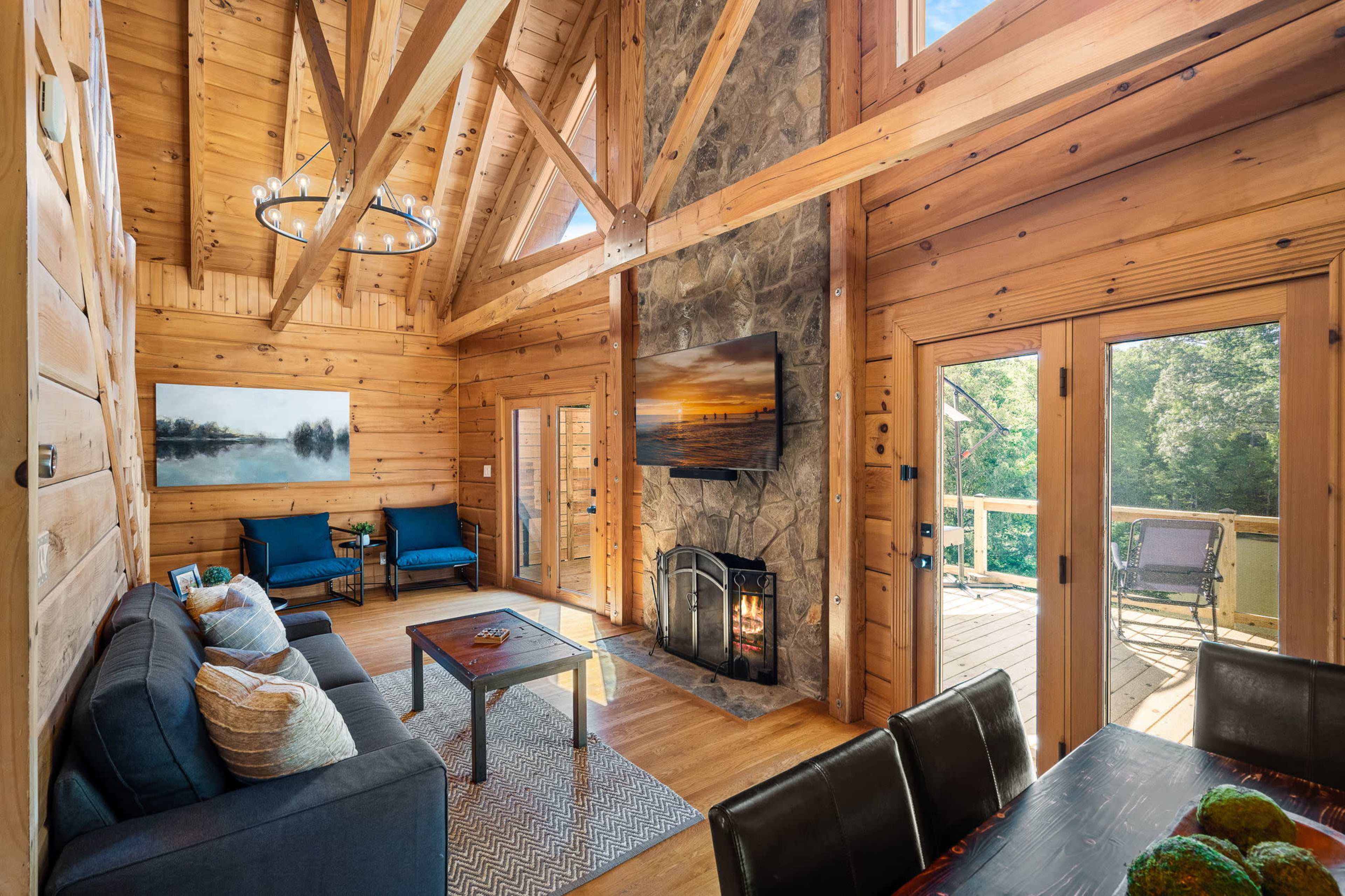 A spacious wooden cabin living room features a stone fireplace, a large window overlooking a deck, and a seating area with a sofa and chairs.