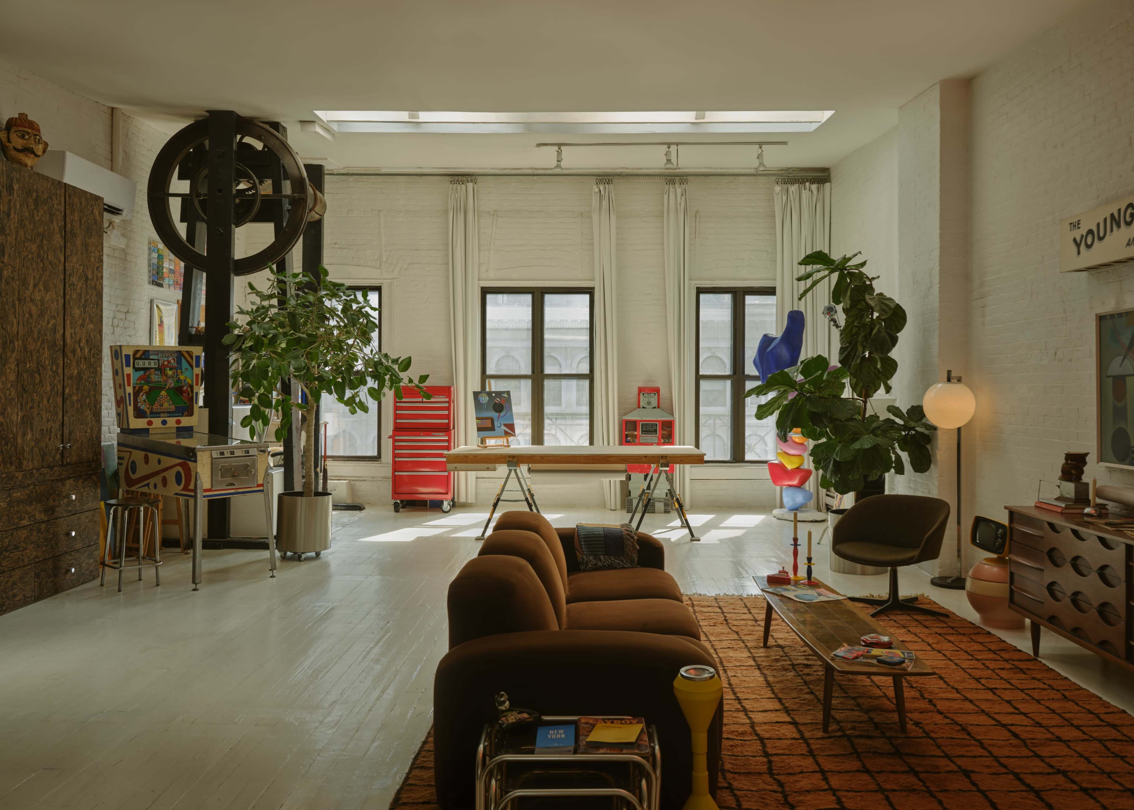 The image shows a spacious, modern living room with large windows, eclectic decor, and a mix of furniture styles including a brown sofa, a coffee table, and various plants.