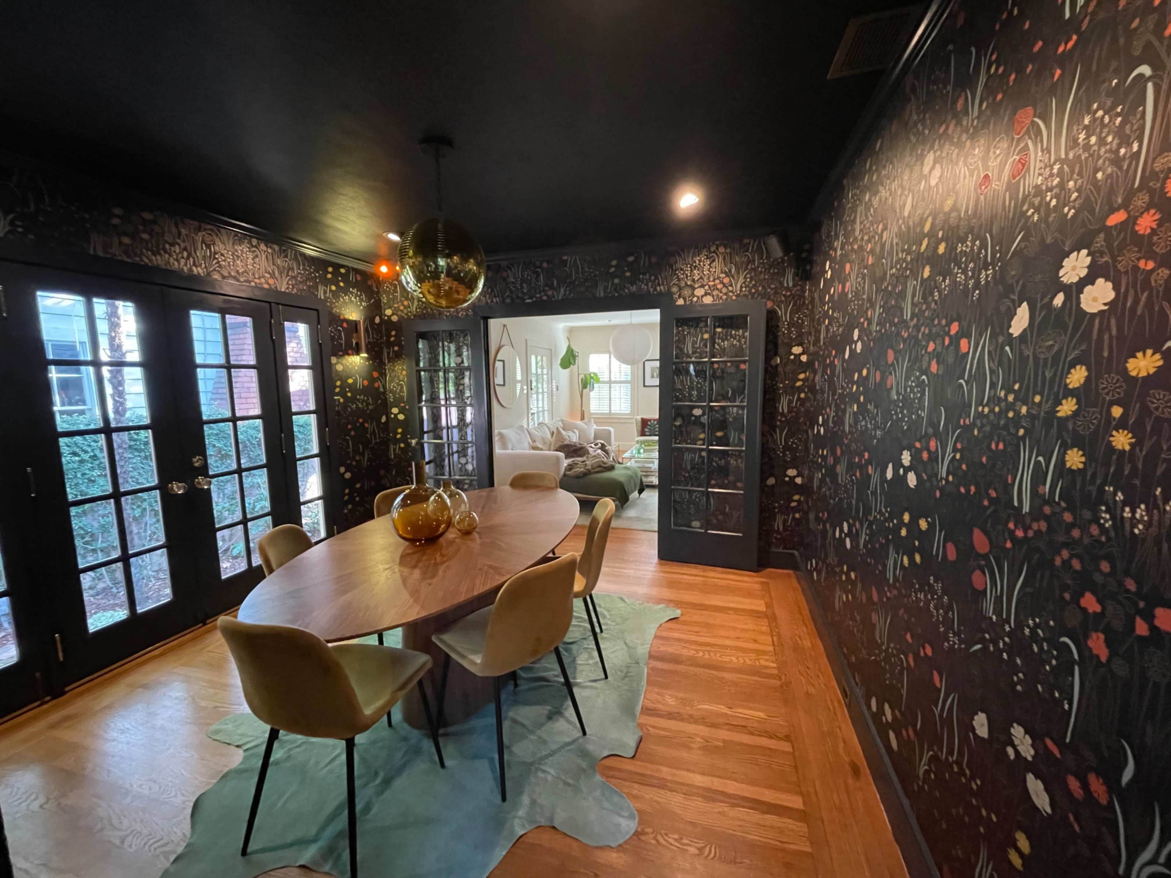 A dining room features a round wooden table with four chairs, surrounded by decorative floral wallpaper and large glass doors leading to another room.