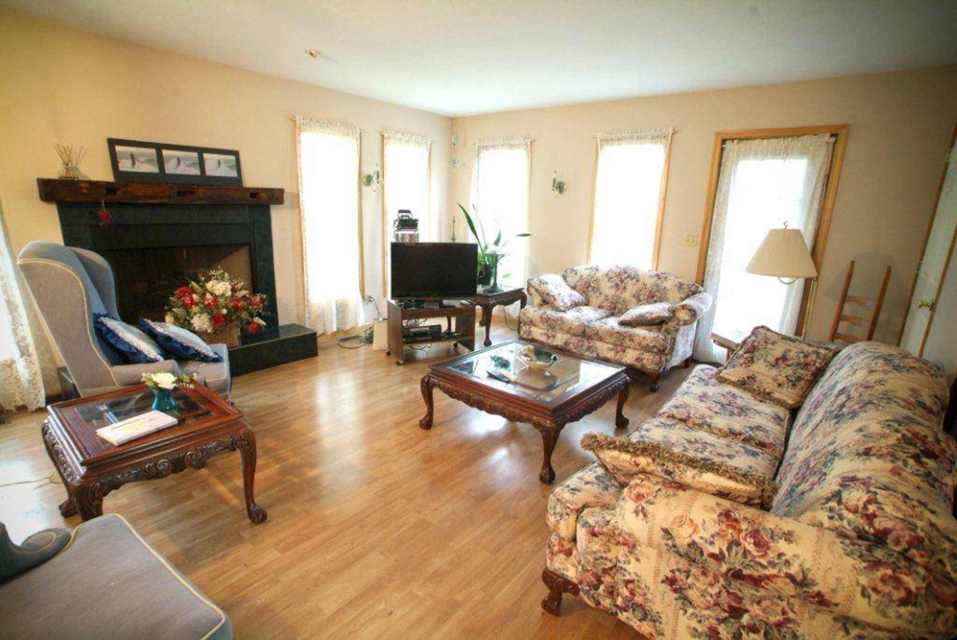 A living room with floral-patterned furniture, a dark fireplace, a television, and large windows allowing natural light.