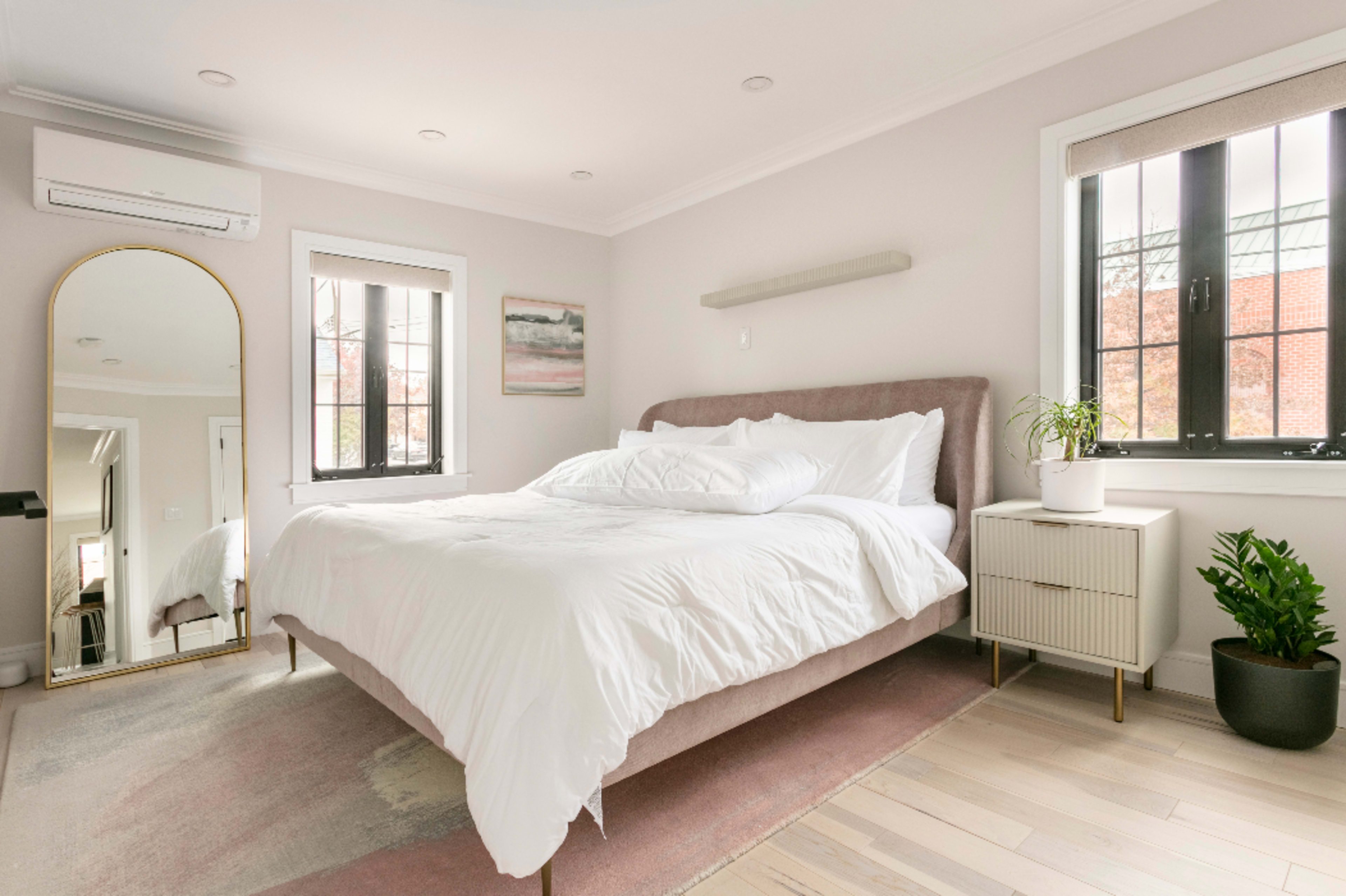 A bright and minimalist bedroom features a king-sized bed with white bedding, a large mirror, a bedside table, and a small potted plant.