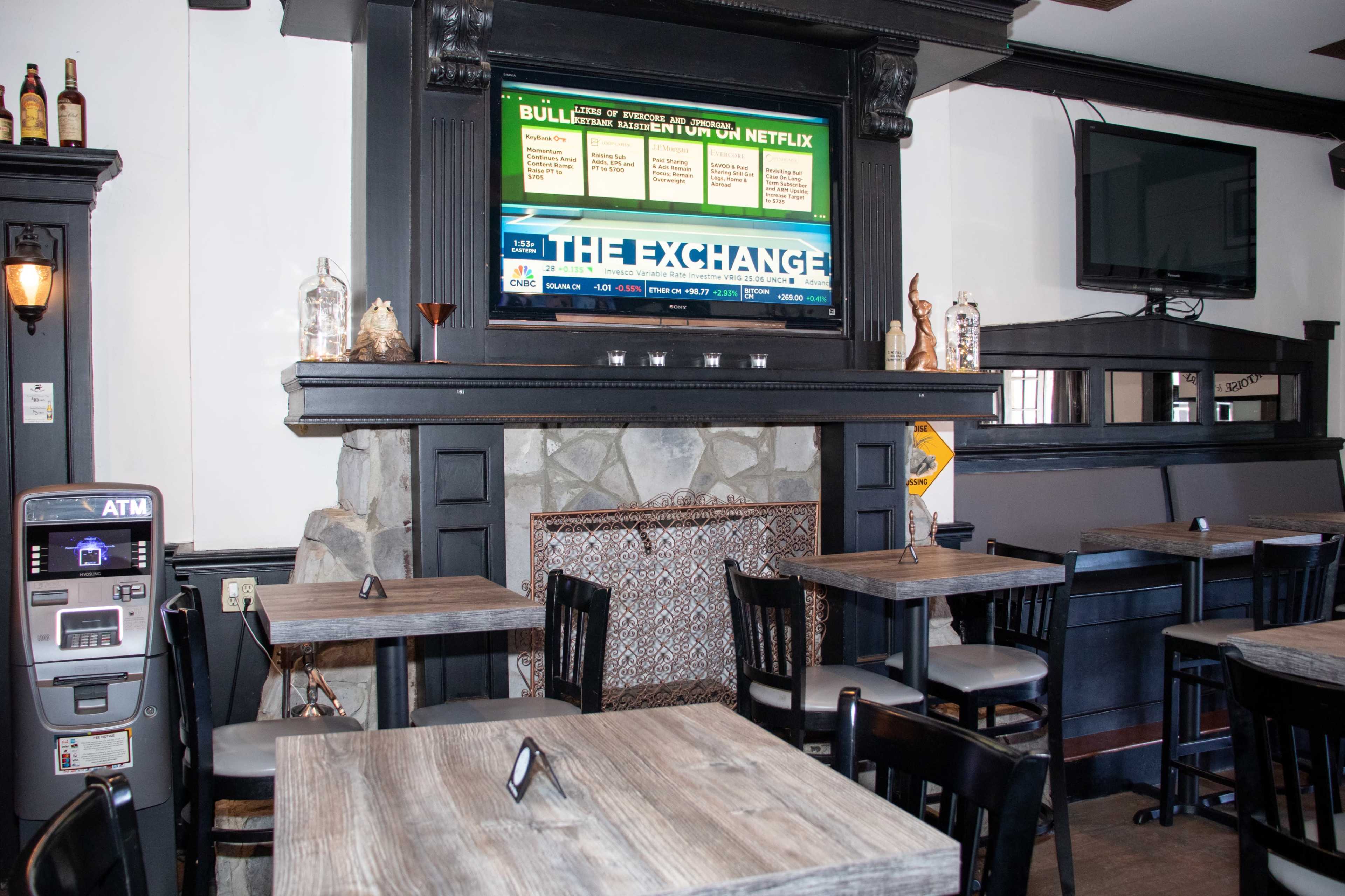 A restaurant interior featuring wooden tables and chairs, a stone fireplace, and a large screen displaying information above the mantel.