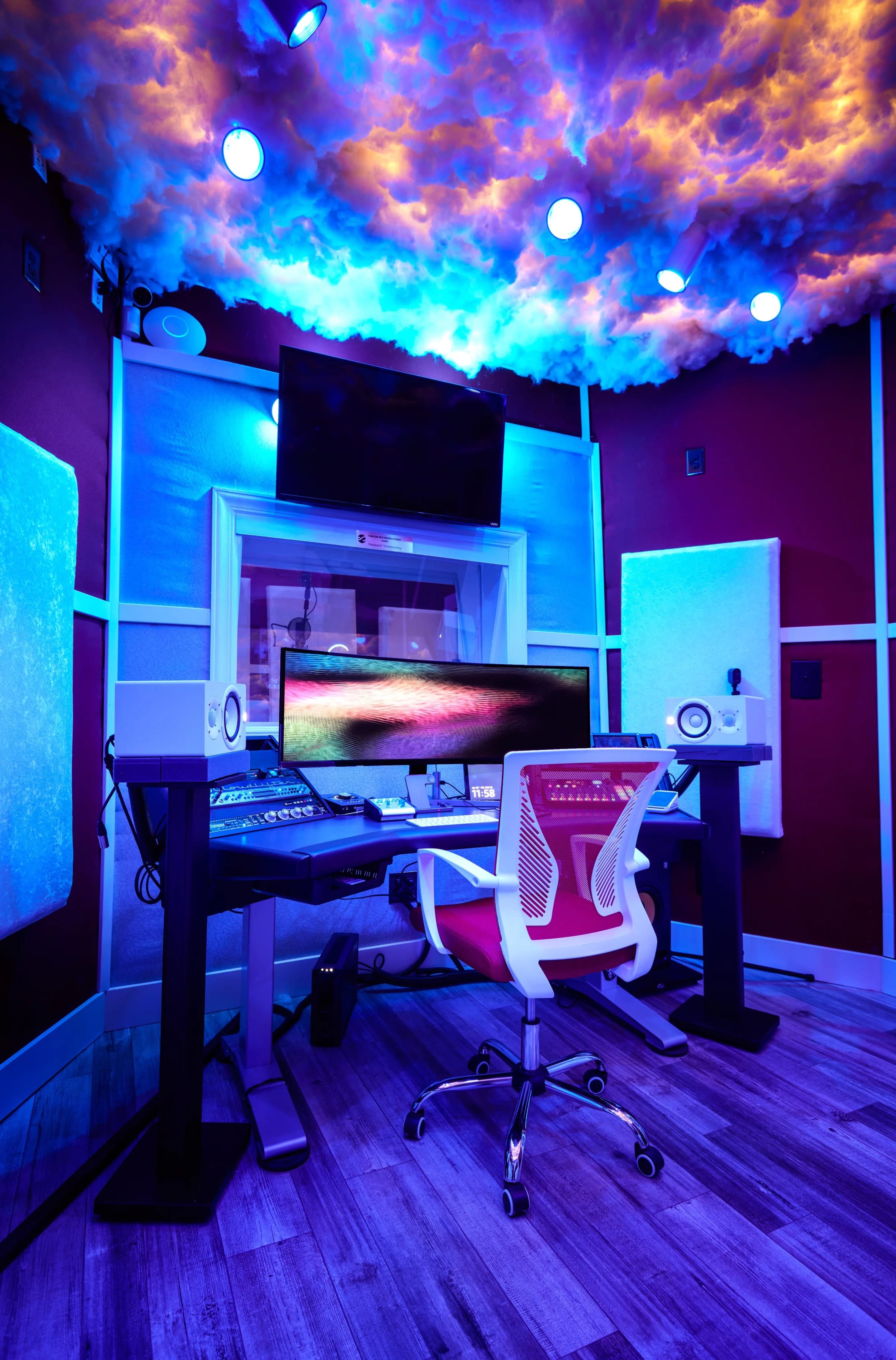 The image shows a modern recording studio featuring a sleek desk with a large monitor, studio monitors, and colorful cloud-like lighting on the ceiling.