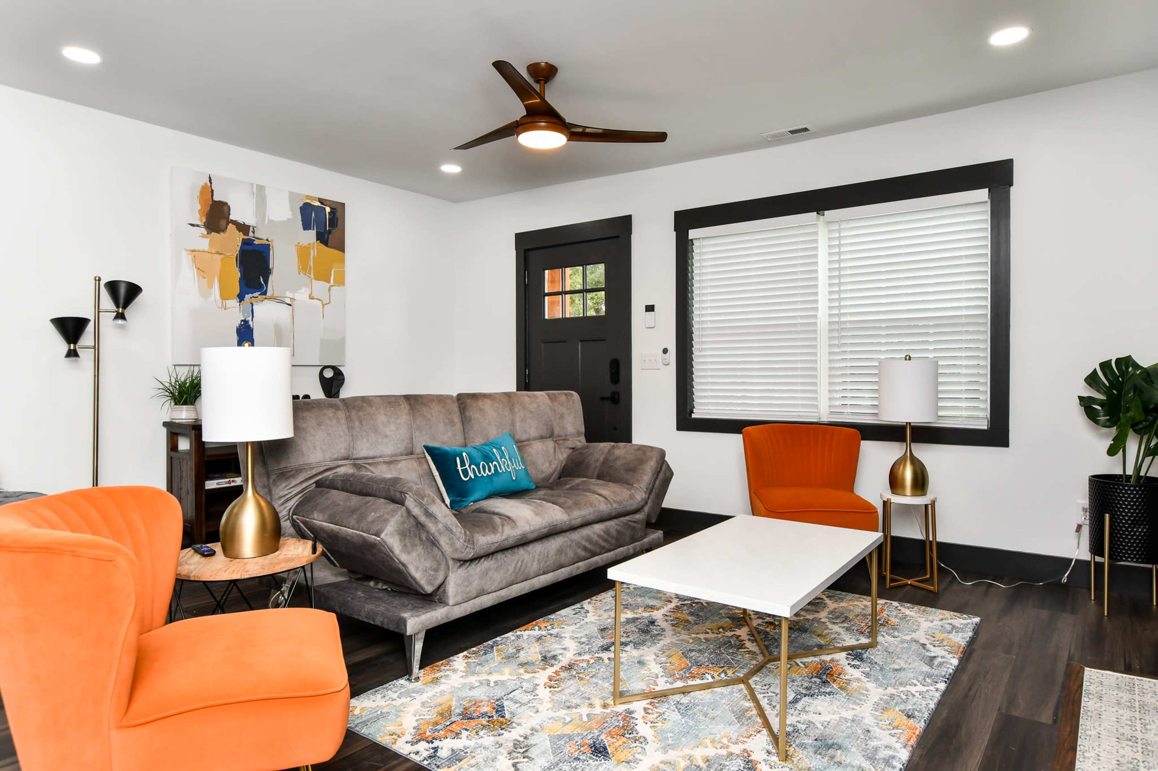 A modern living room with a gray sofa, two orange armchairs, a white coffee table on a patterned rug, and a large piece of abstract art on the wall.