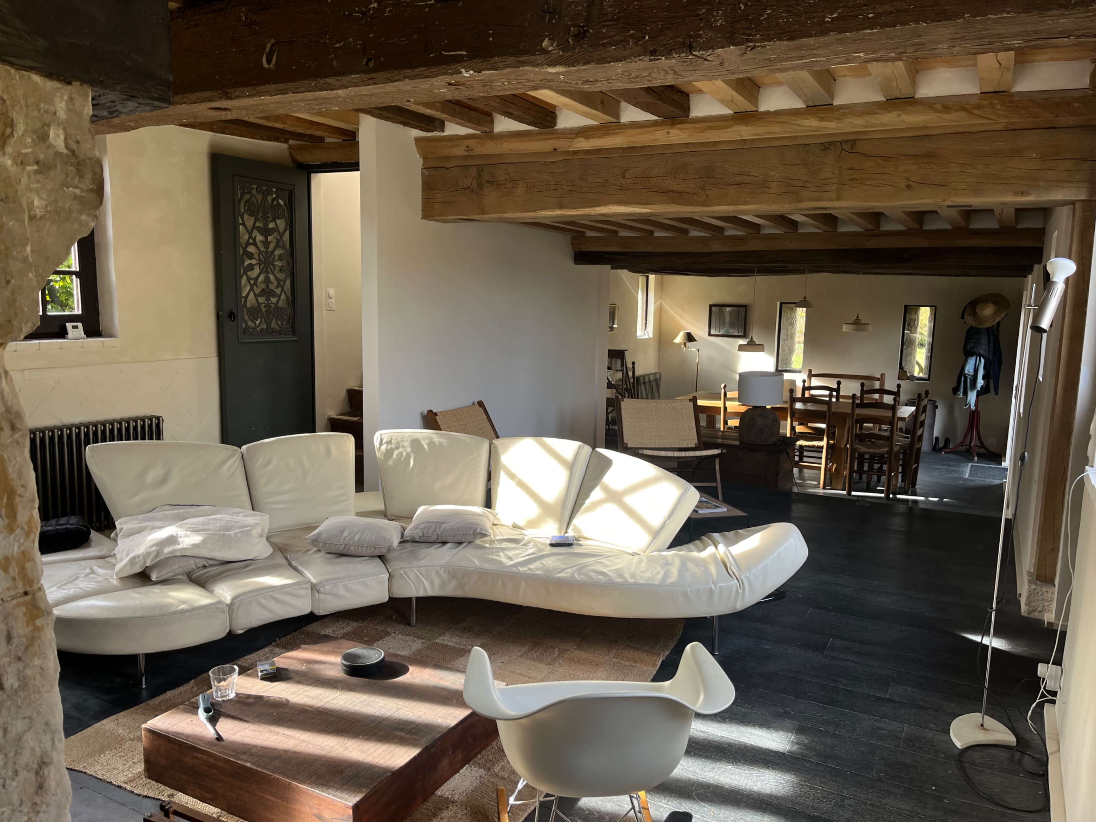 The image shows a modern living space with a curved white sofa, a wooden coffee table, and a dining area in the background featuring wooden furniture.
