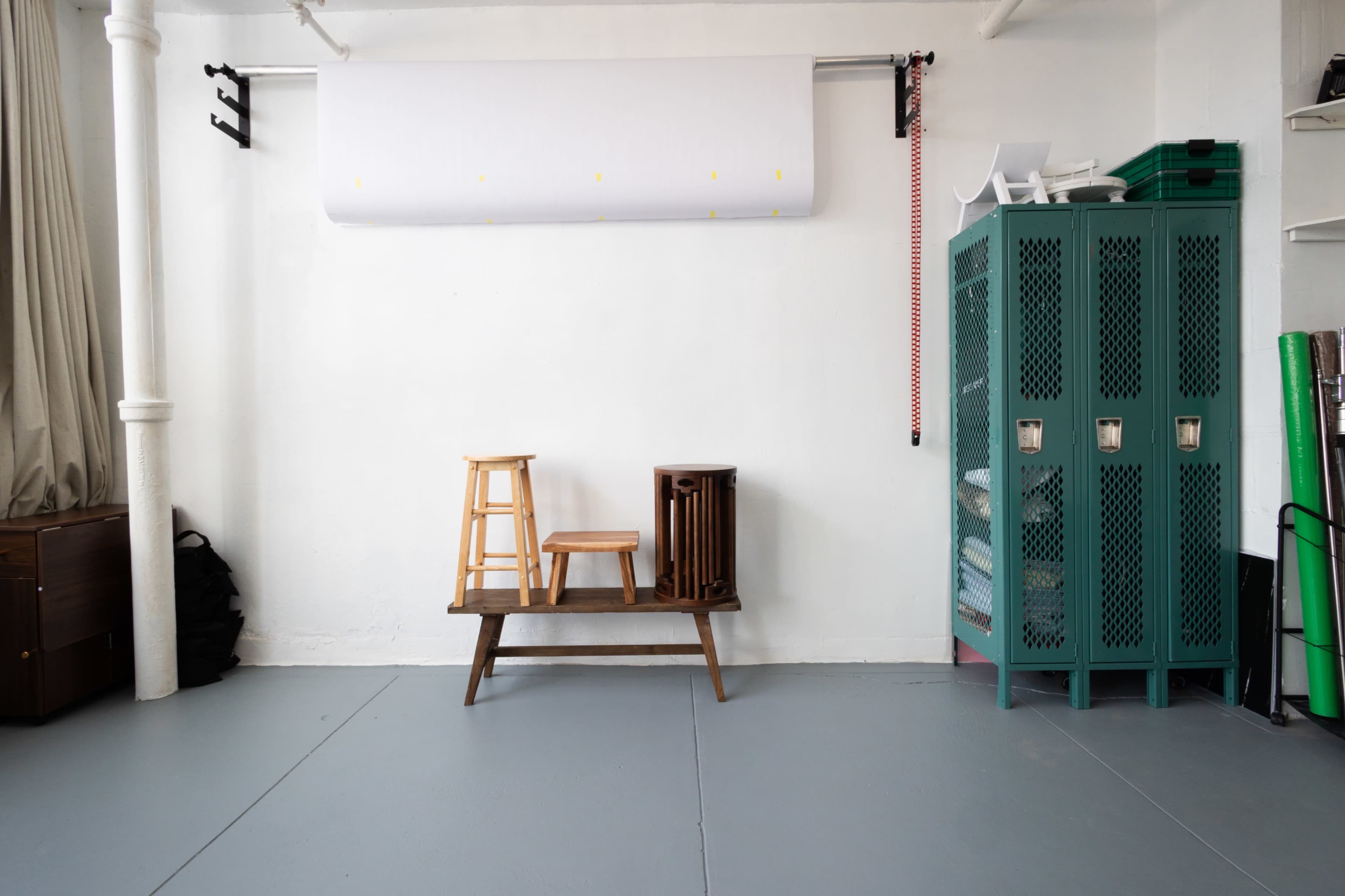 The image shows a minimalistic studio space with a light-colored wall, a paper roll mounted above, two wooden stools placed on a wooden bench, and a green storage unit beside rolled-up materials.