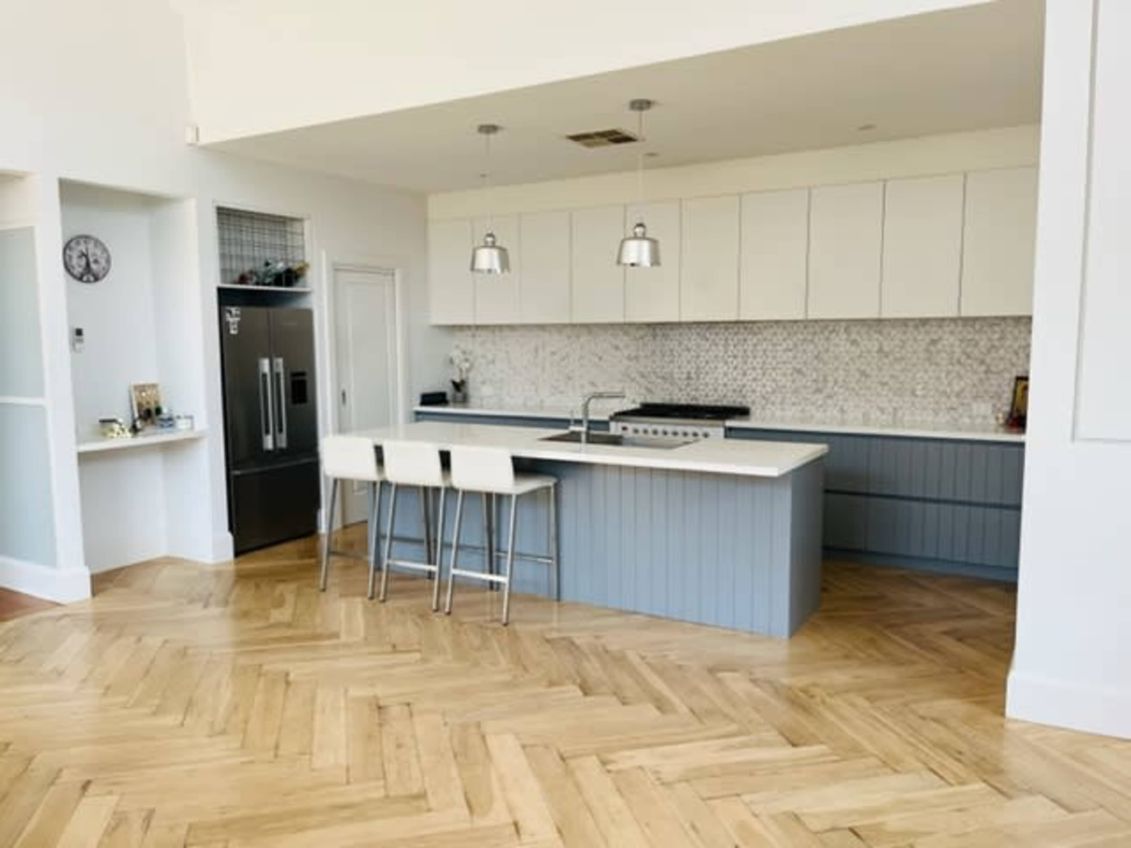 The image shows a modern kitchen featuring a large island with bar stools, stainless steel appliances, and a herringbone wood floor.