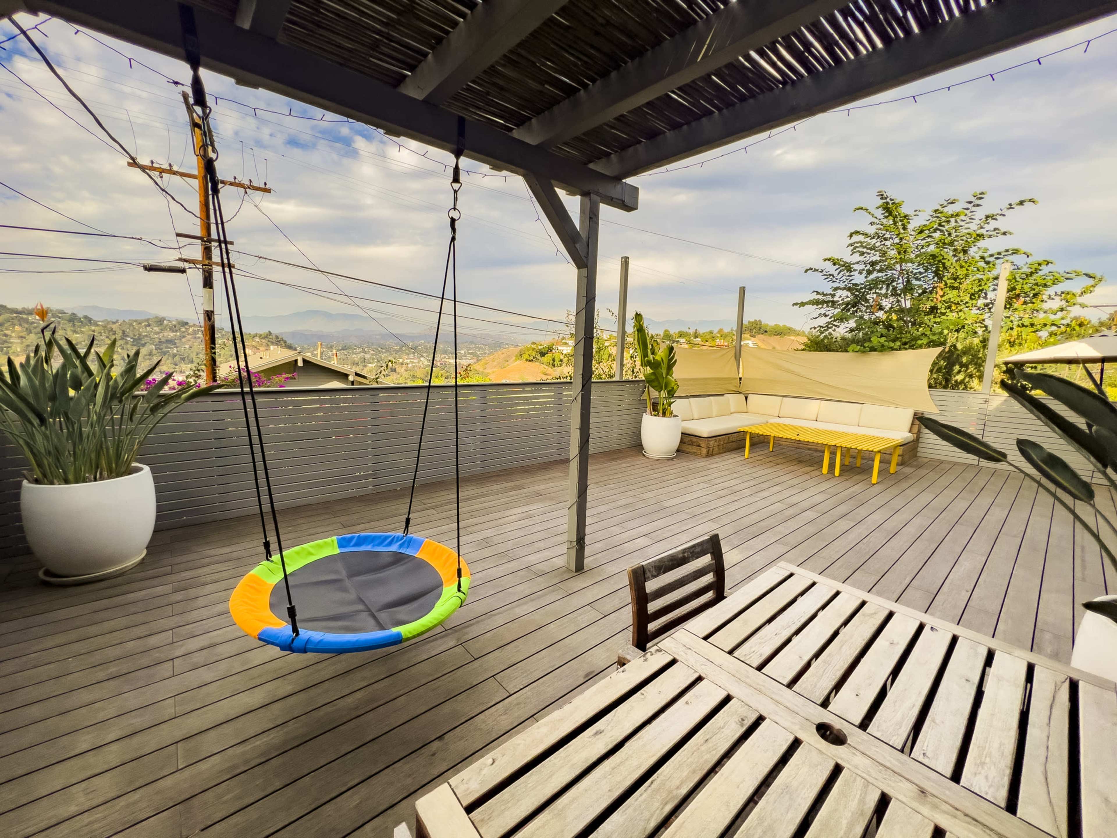 A wooden deck features a colorful swing, a dining table, and surrounding potted plants with a view of rolling hills in the background.