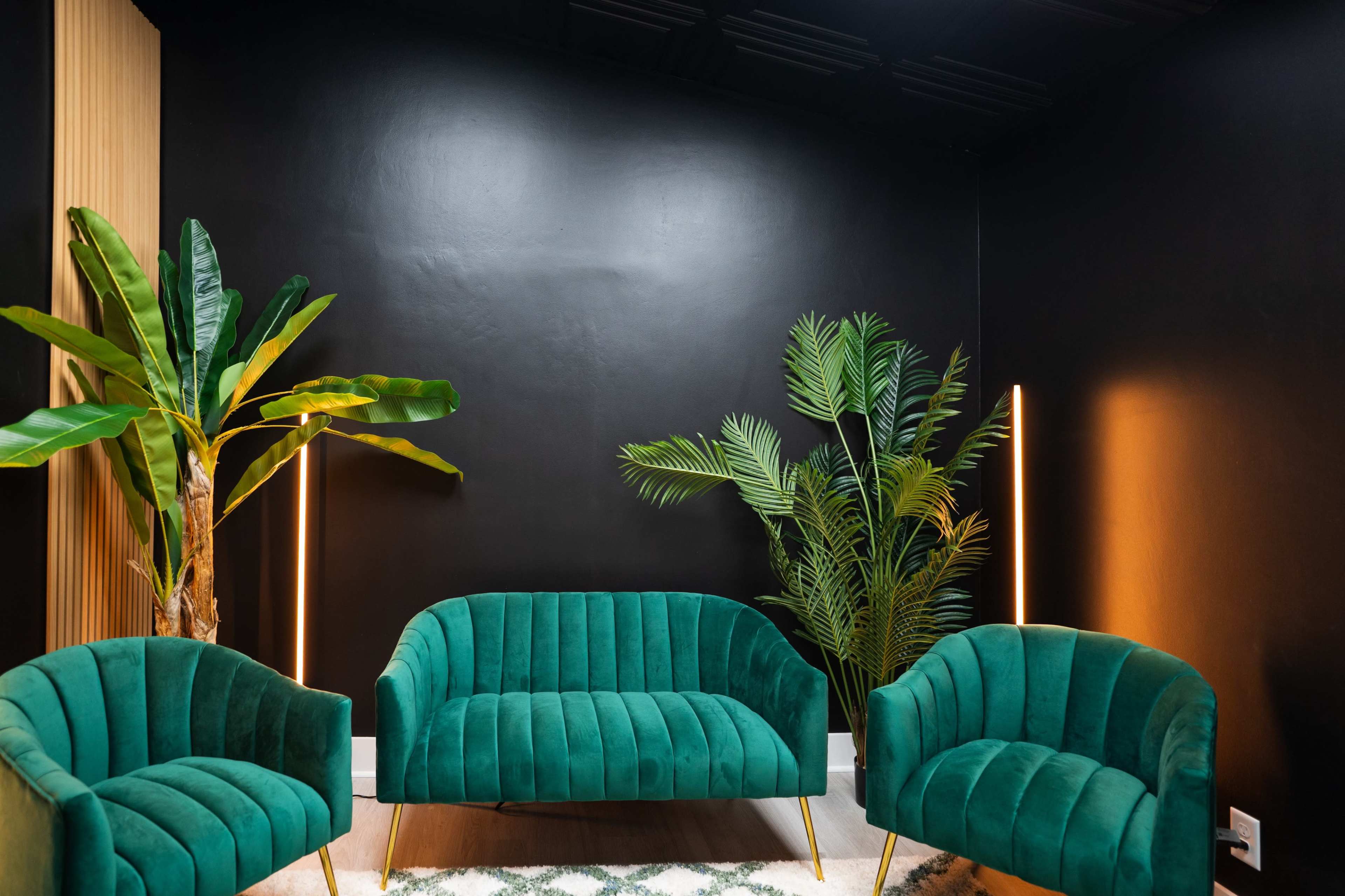 A modern seating area features three green velvet chairs and several indoor plants against a black wall.