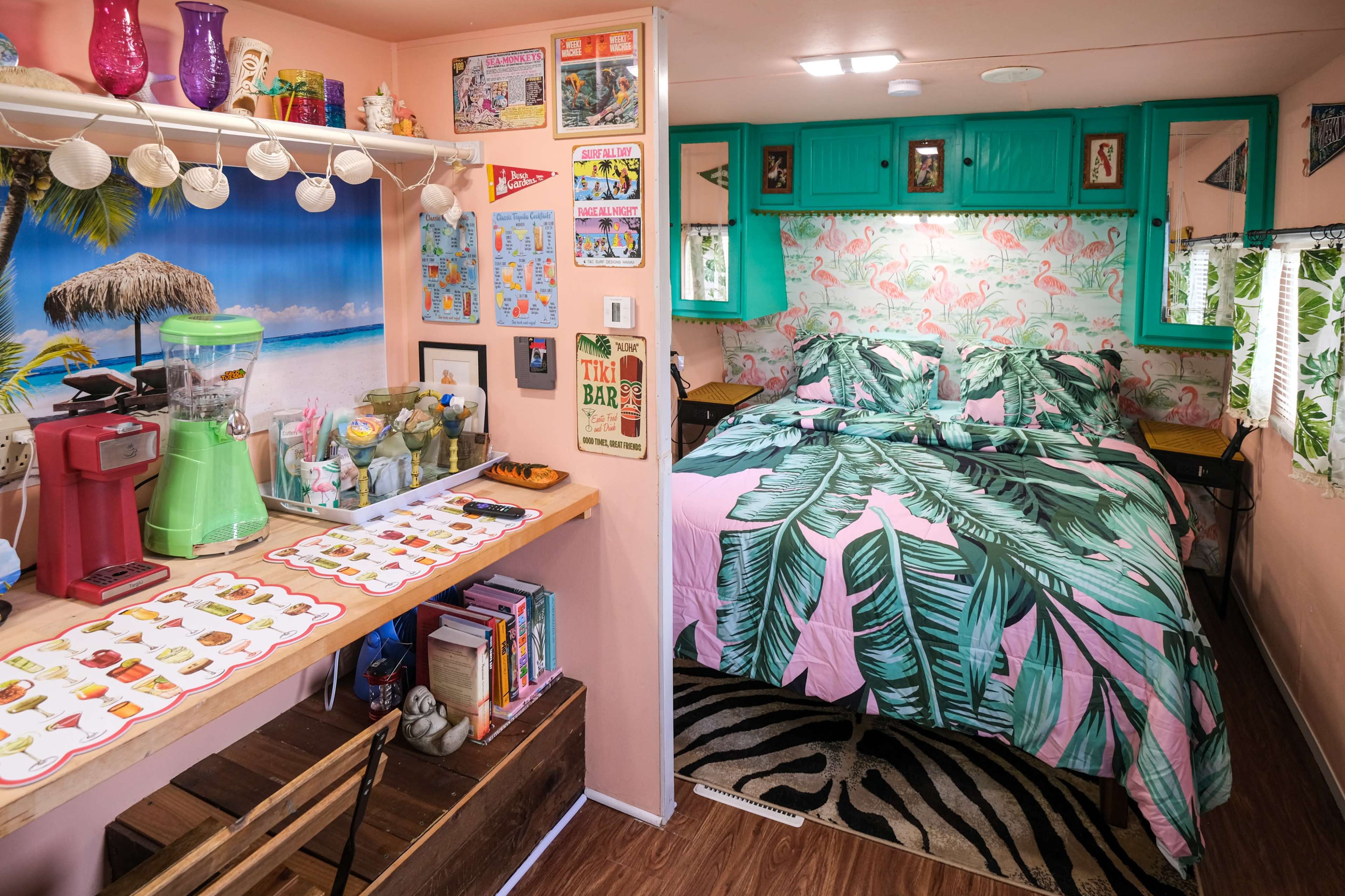 The image features a small, colorful living space that combines a kitchen area with a dining table and a bedroom, adorned with tropical-themed decor and vibrant palm leaf patterns.