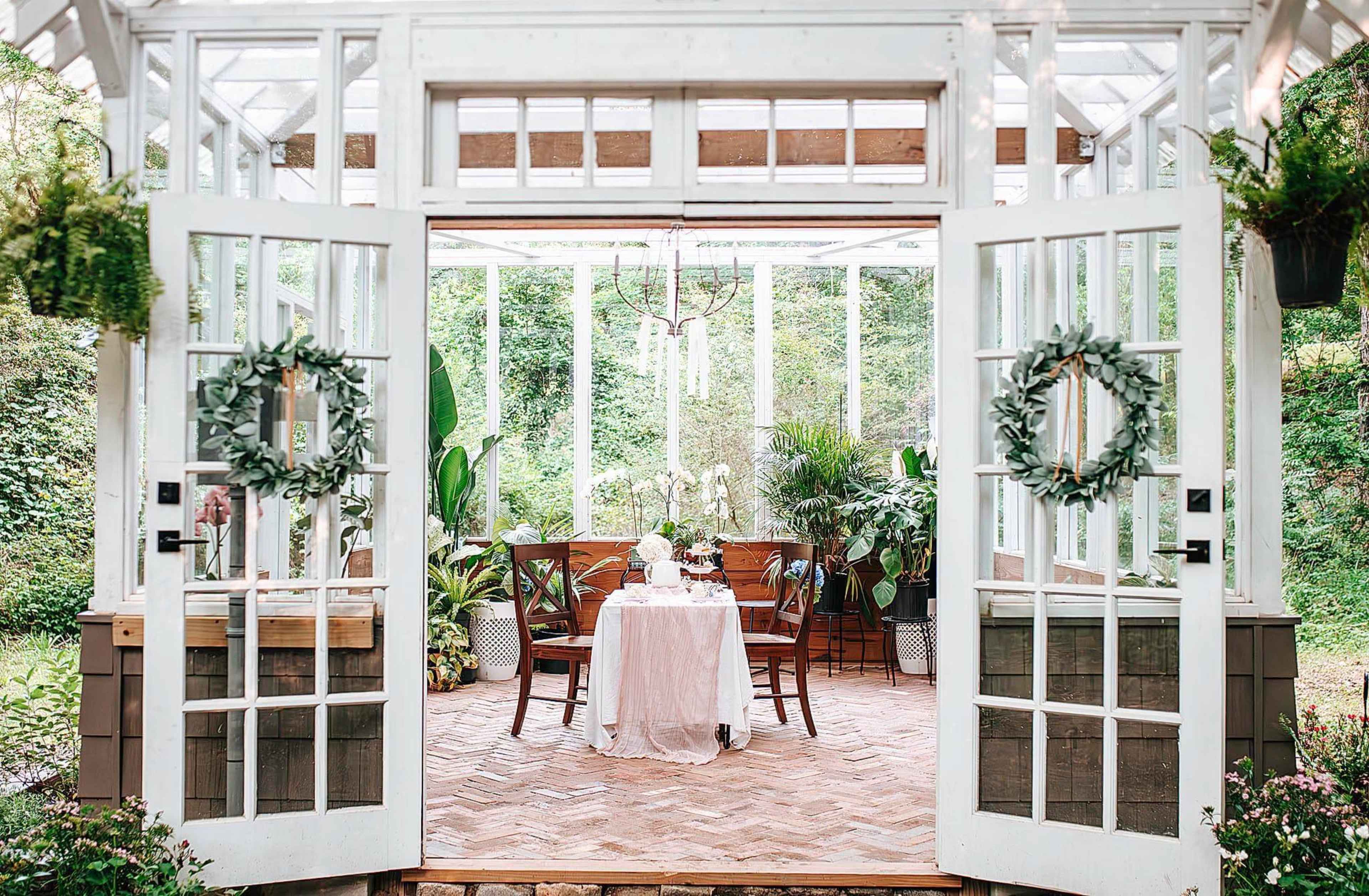 A glass-enclosed garden room features a circular dining table set for two, surrounded by lush greenery and decorative plants.