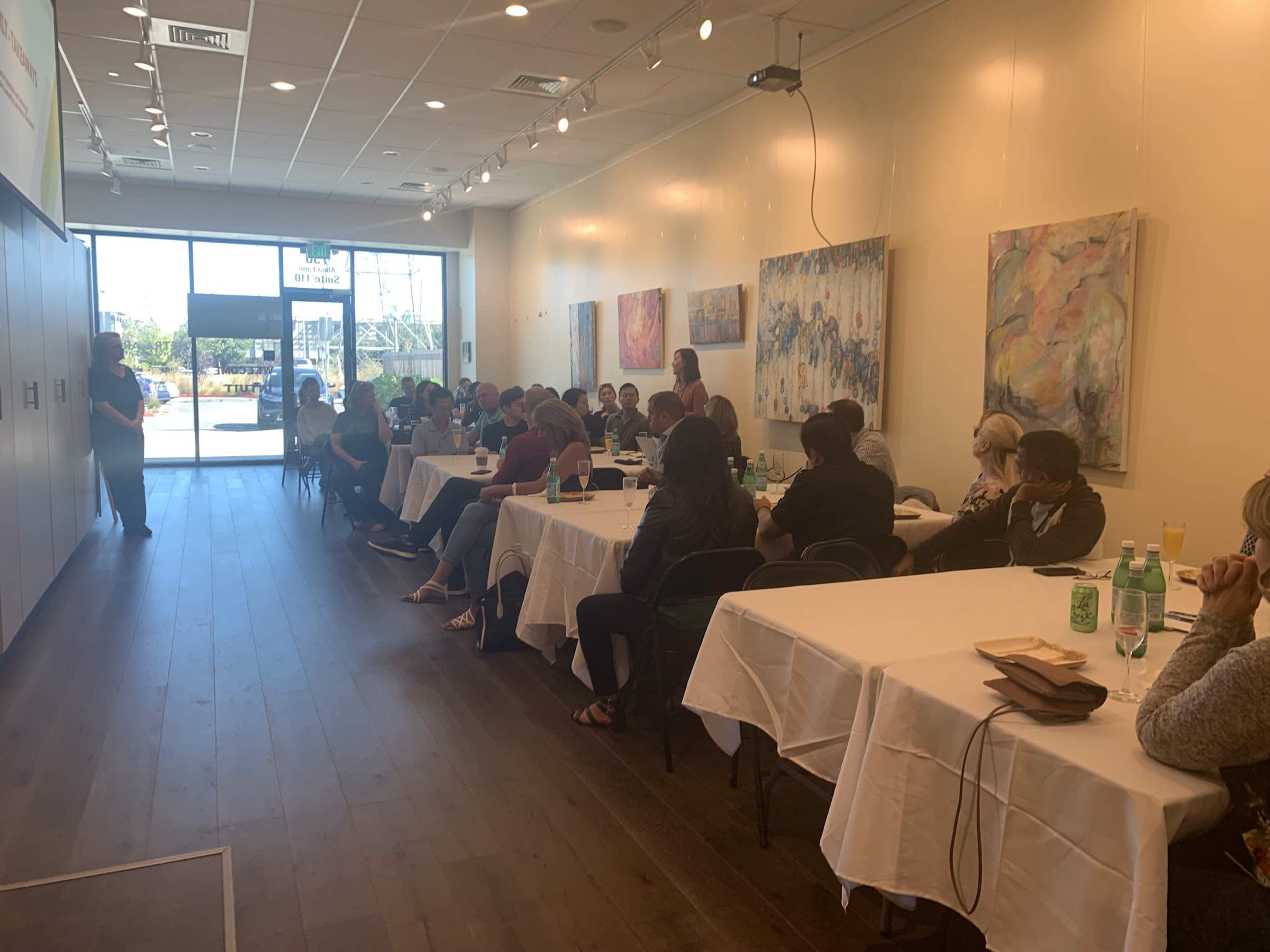 A group of people attentively sits at long tables in a brightly lit room with artworks on the walls.