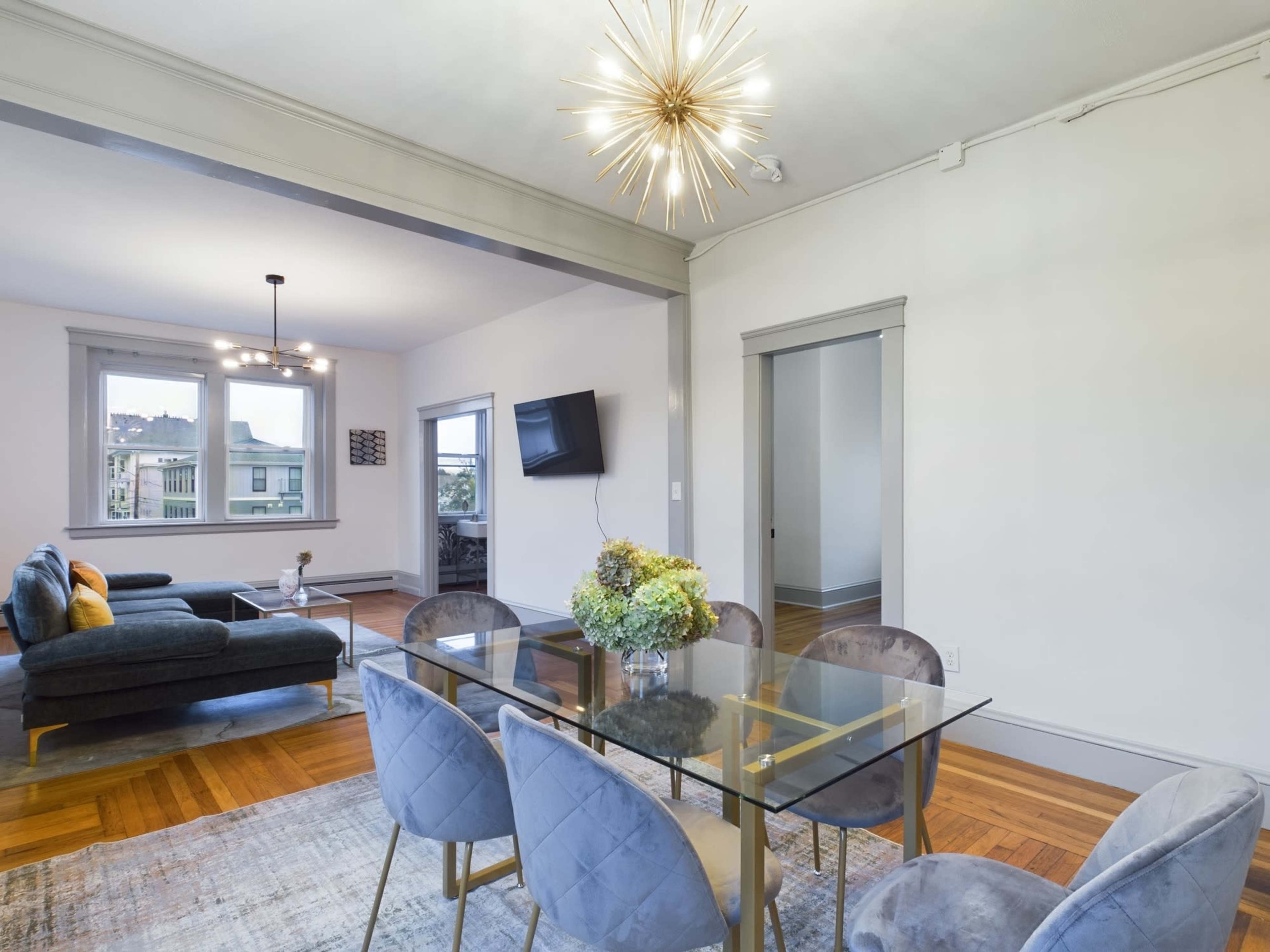 A modern dining area with a glass table surrounded by upholstered chairs and a decorative light fixture overhead, adjacent to a living space with a sofa.