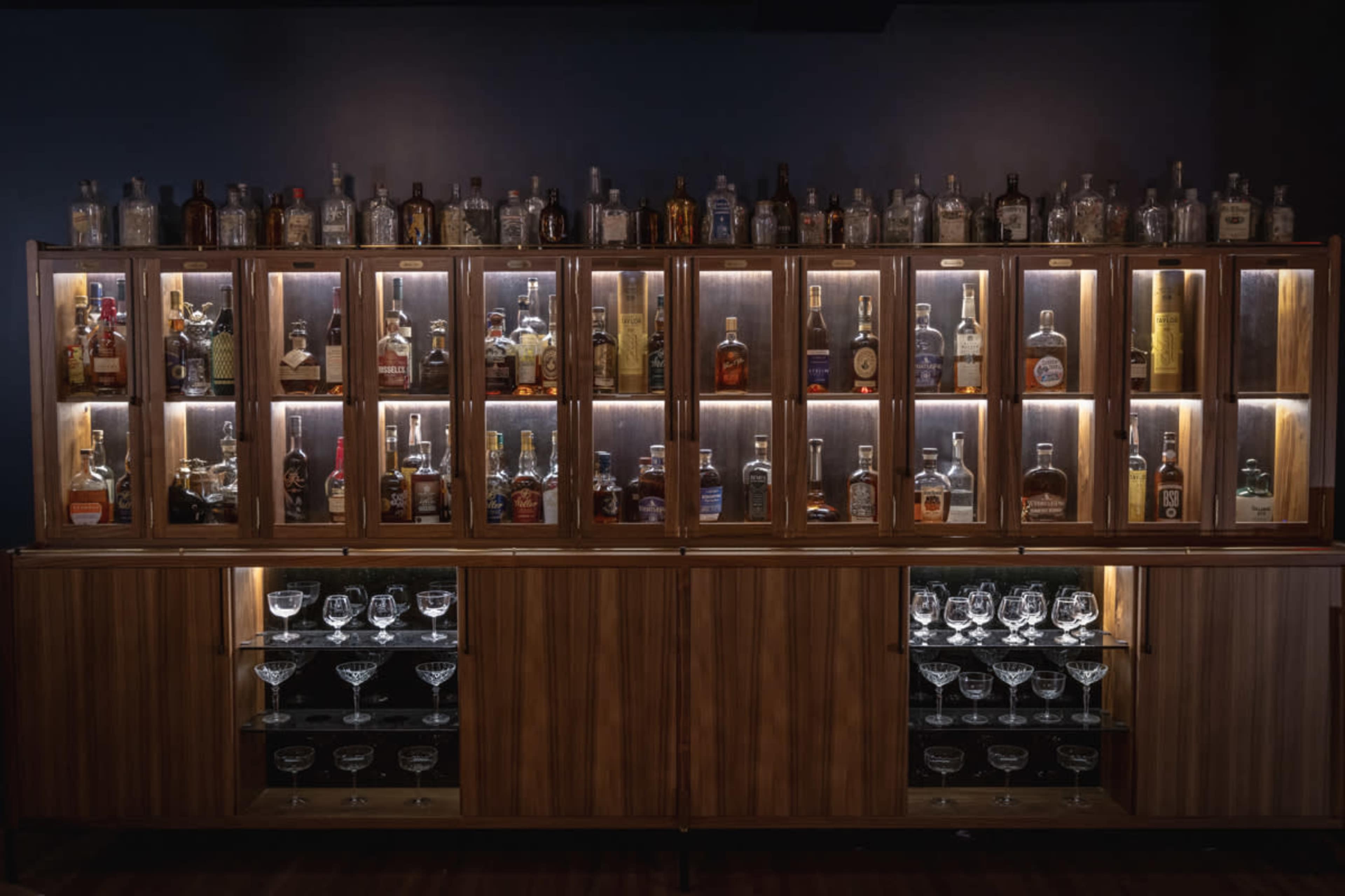 A wooden bar cabinet filled with various bottles of liquor, illuminated by lights, with a set of glasses displayed underneath.