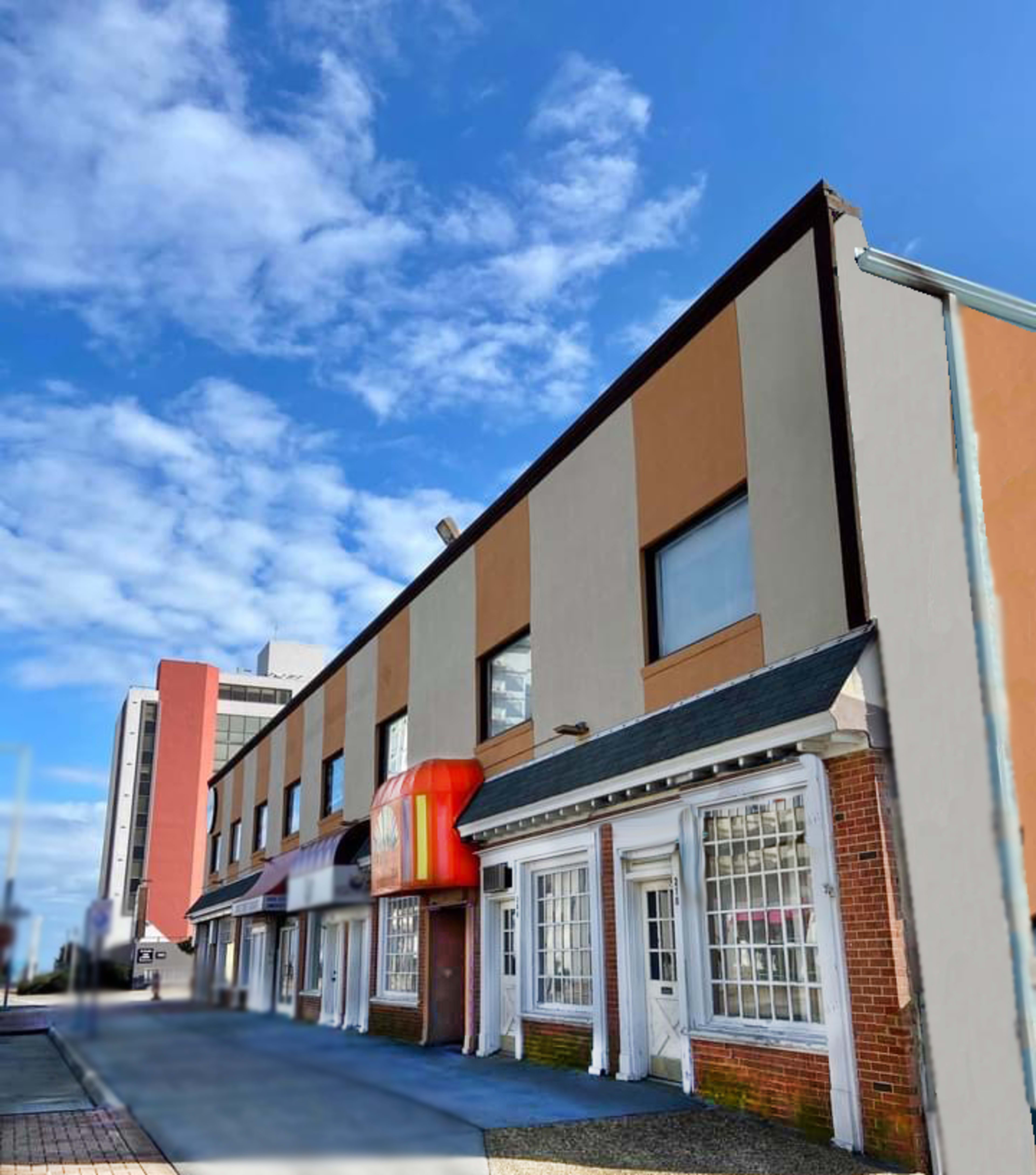 A two-story building with a mix of brown and beige exterior walls and large windows on a clear day, with a partial view of a nearby taller building in the background.