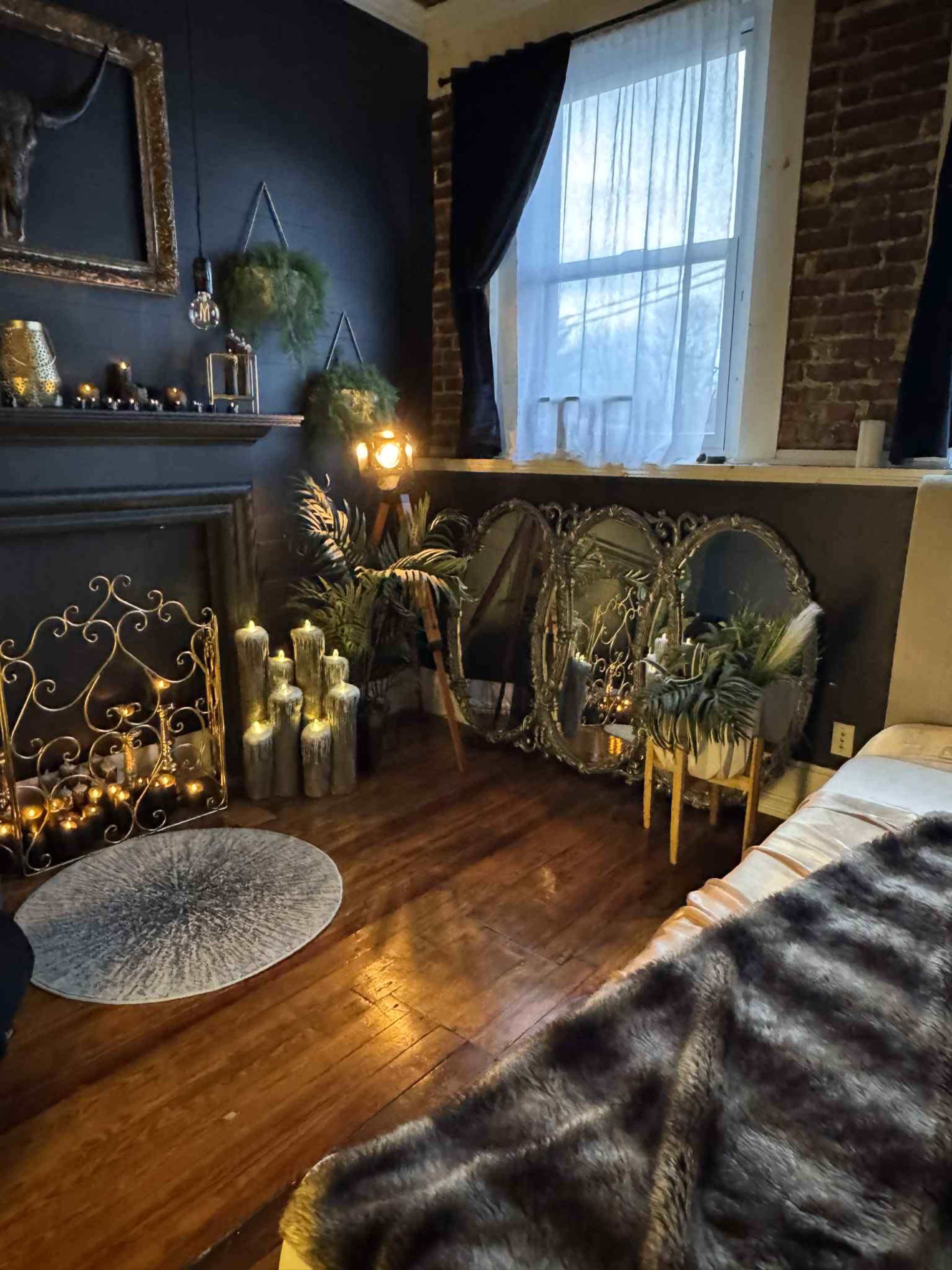 The image shows a cozy interior space with a dark wall, a fireplace adorned with decorative items, mirrors, and plants, illuminated by soft lighting.