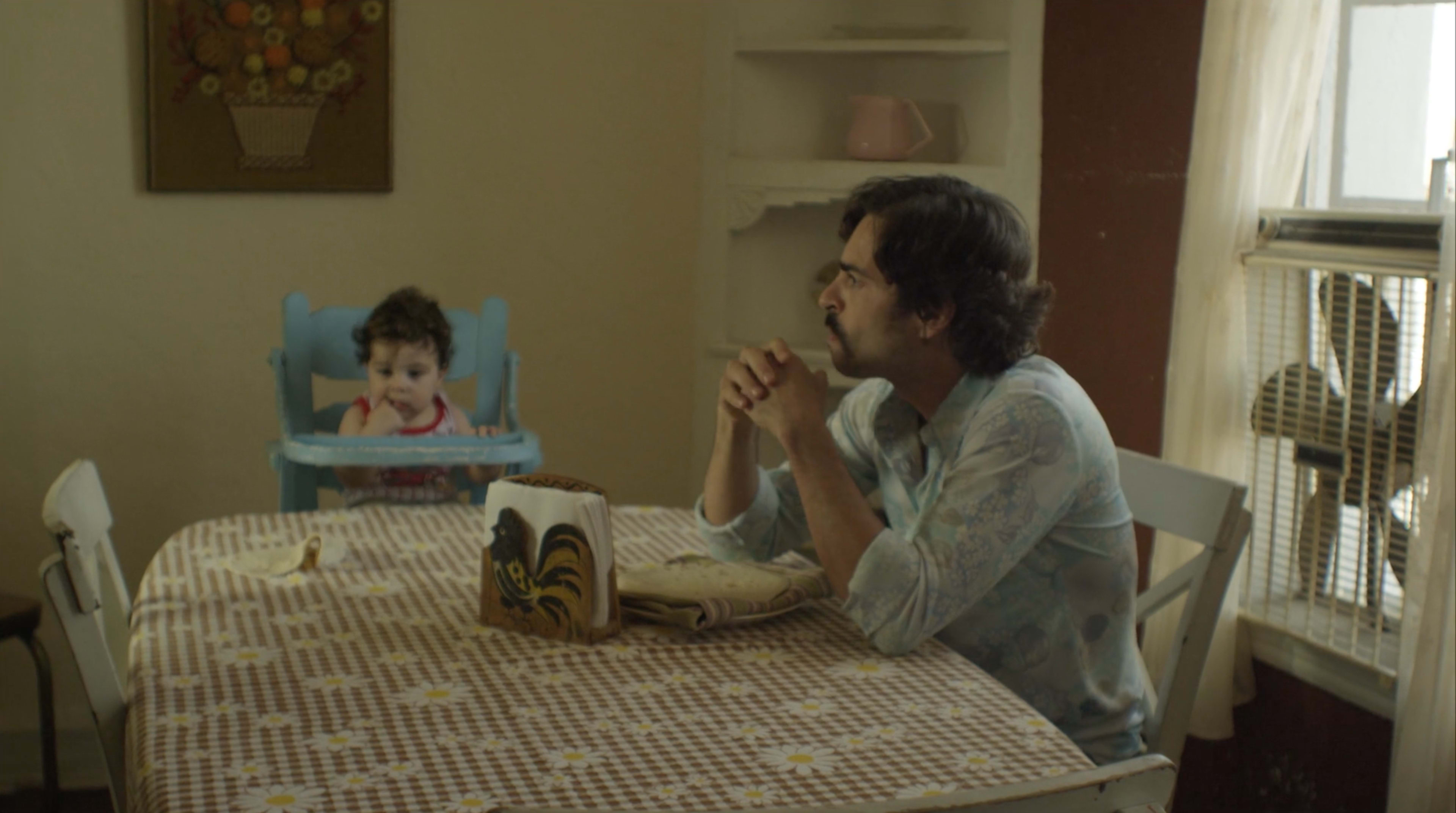 A man sits at a table in a room with a child in a high chair nearby.