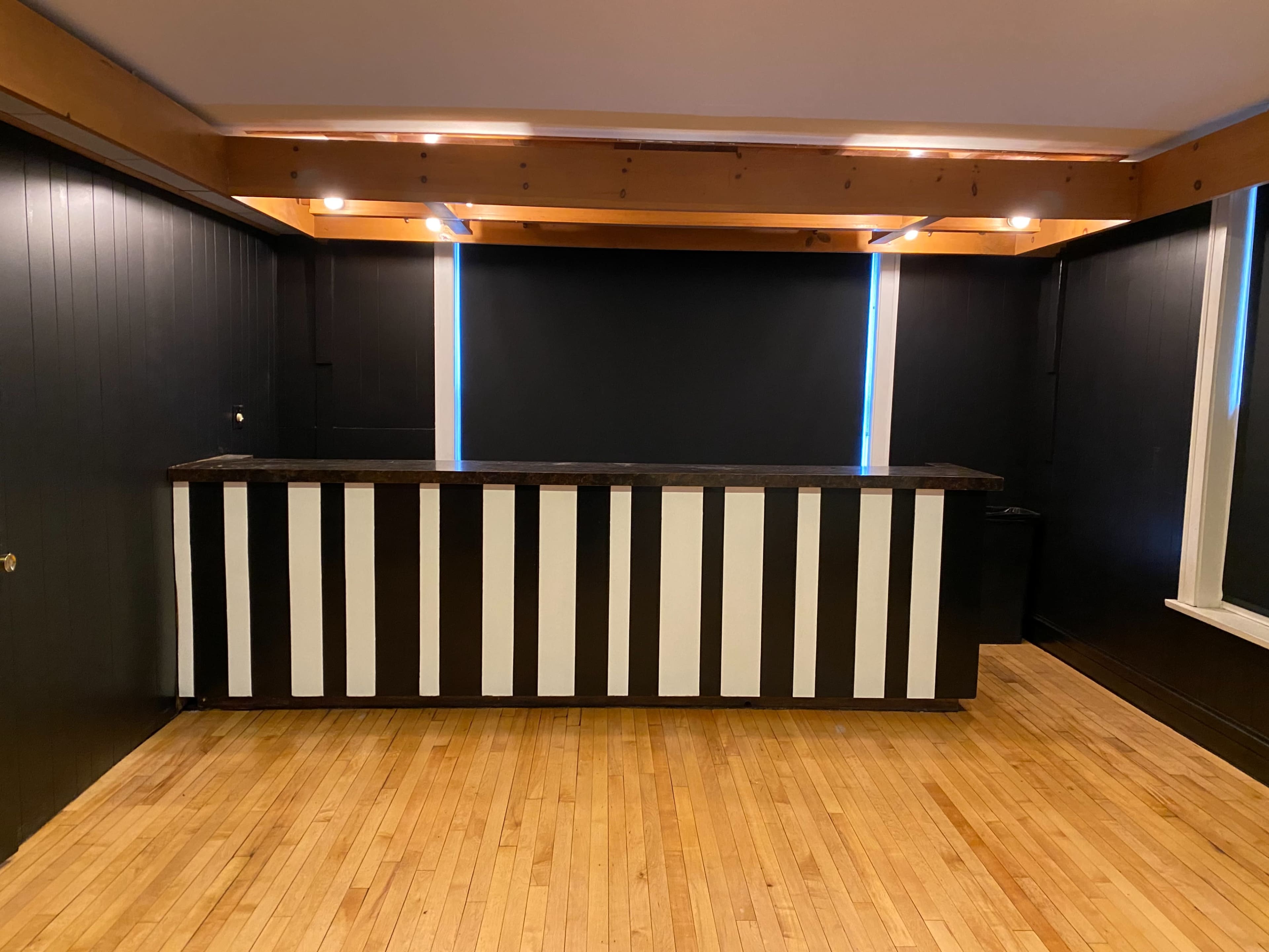A room with a wooden floor, featuring a black and white striped bar counter against dark walls and ceiling beams.