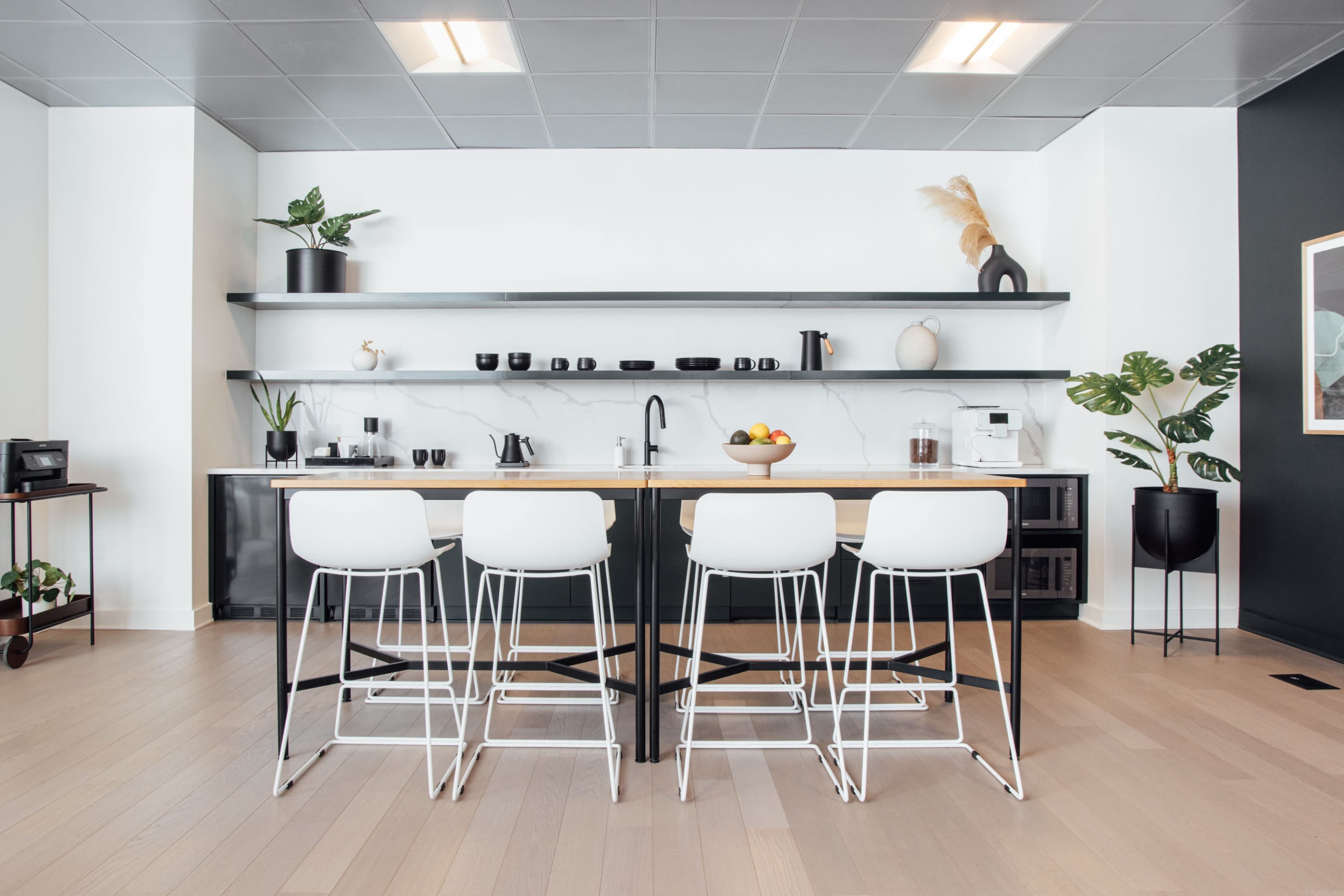 A modern kitchen features a long white counter with four high stools, shelving above displaying various kitchen items, and plants placed throughout the space.