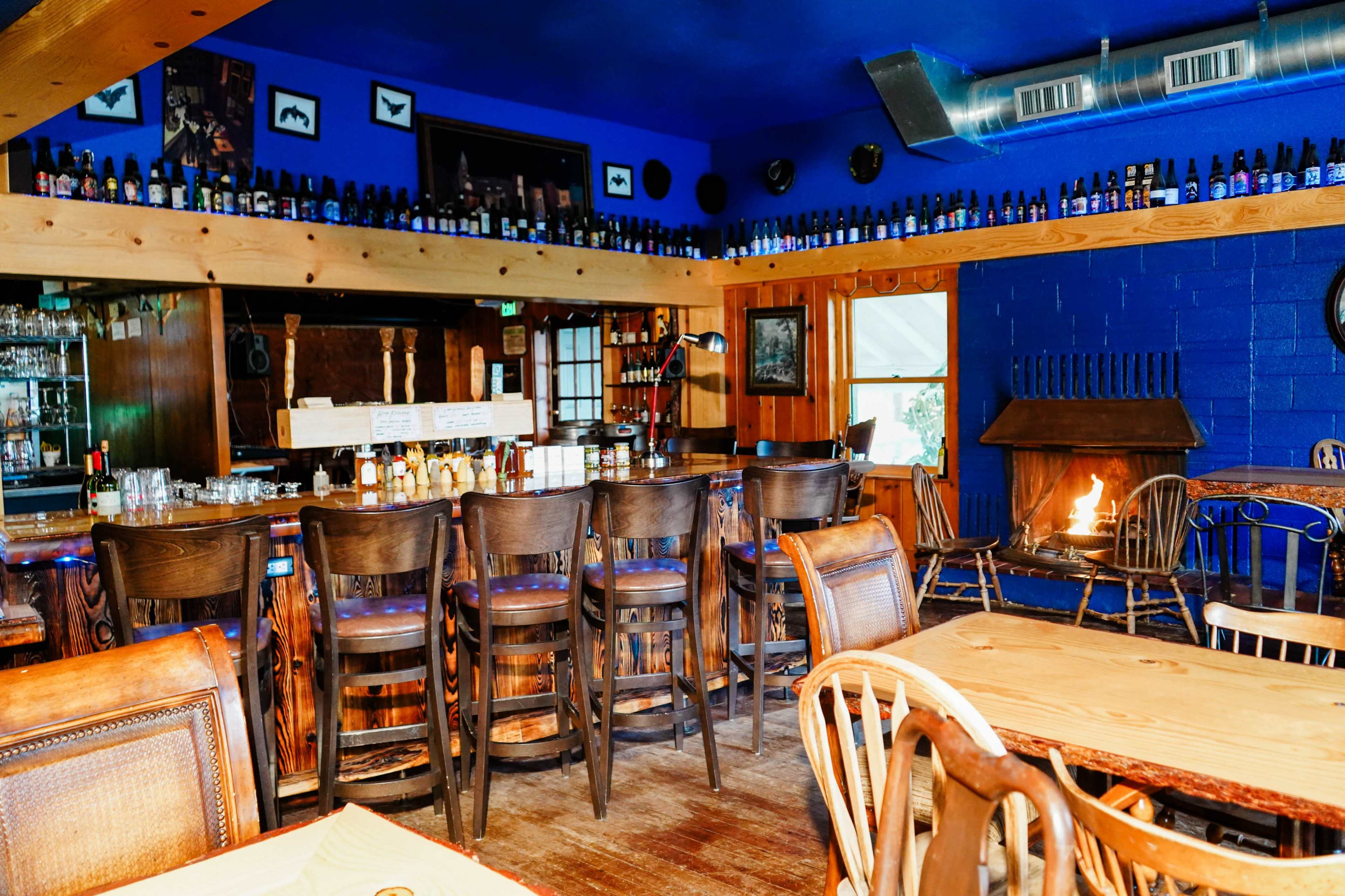 A cozy bar area with wooden chairs, a wooden counter, and a fireplace, all set against blue walls adorned with bottles.