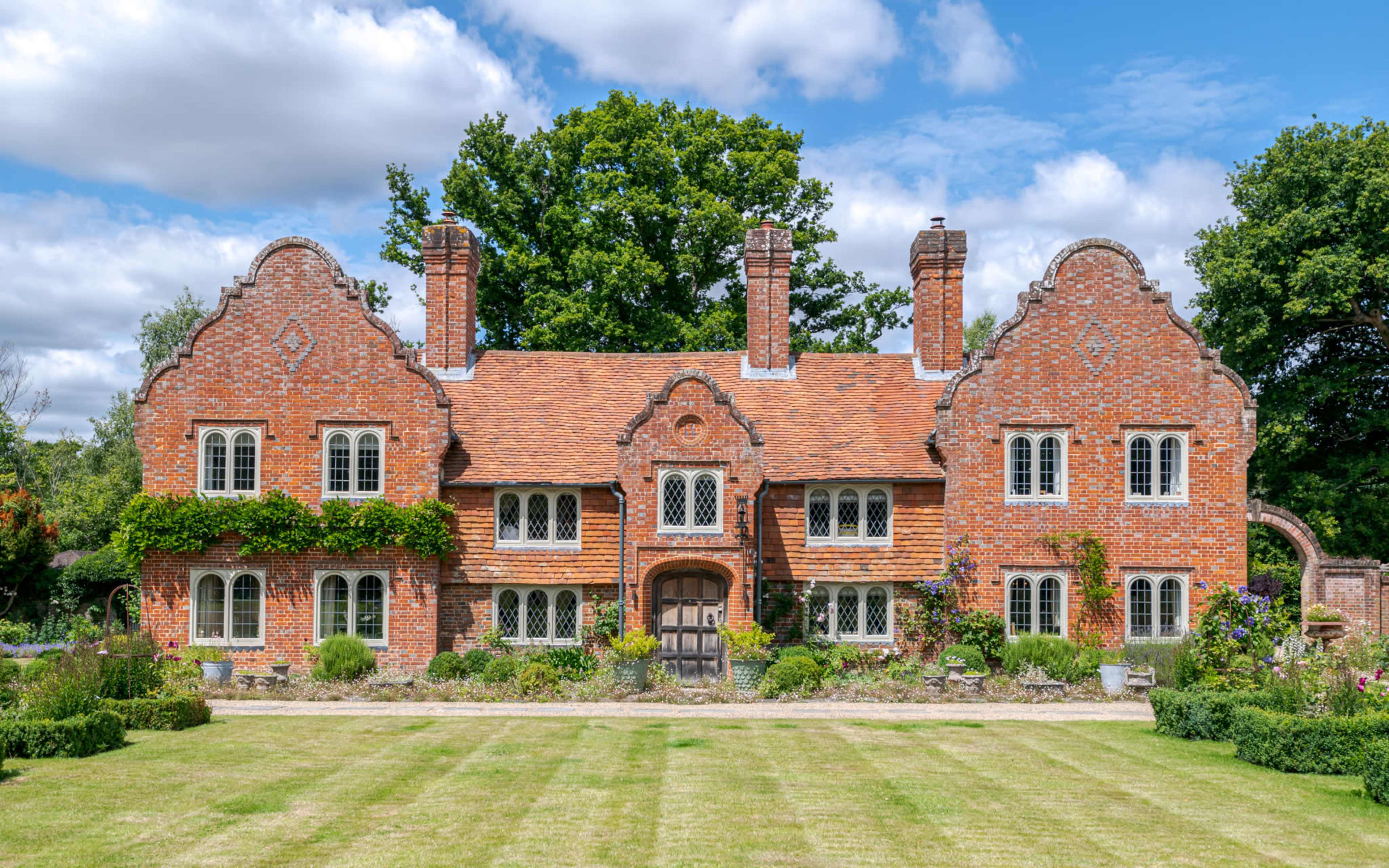 A large, red-brick house with ornate gables and a landscaped garden is set against a blue sky.