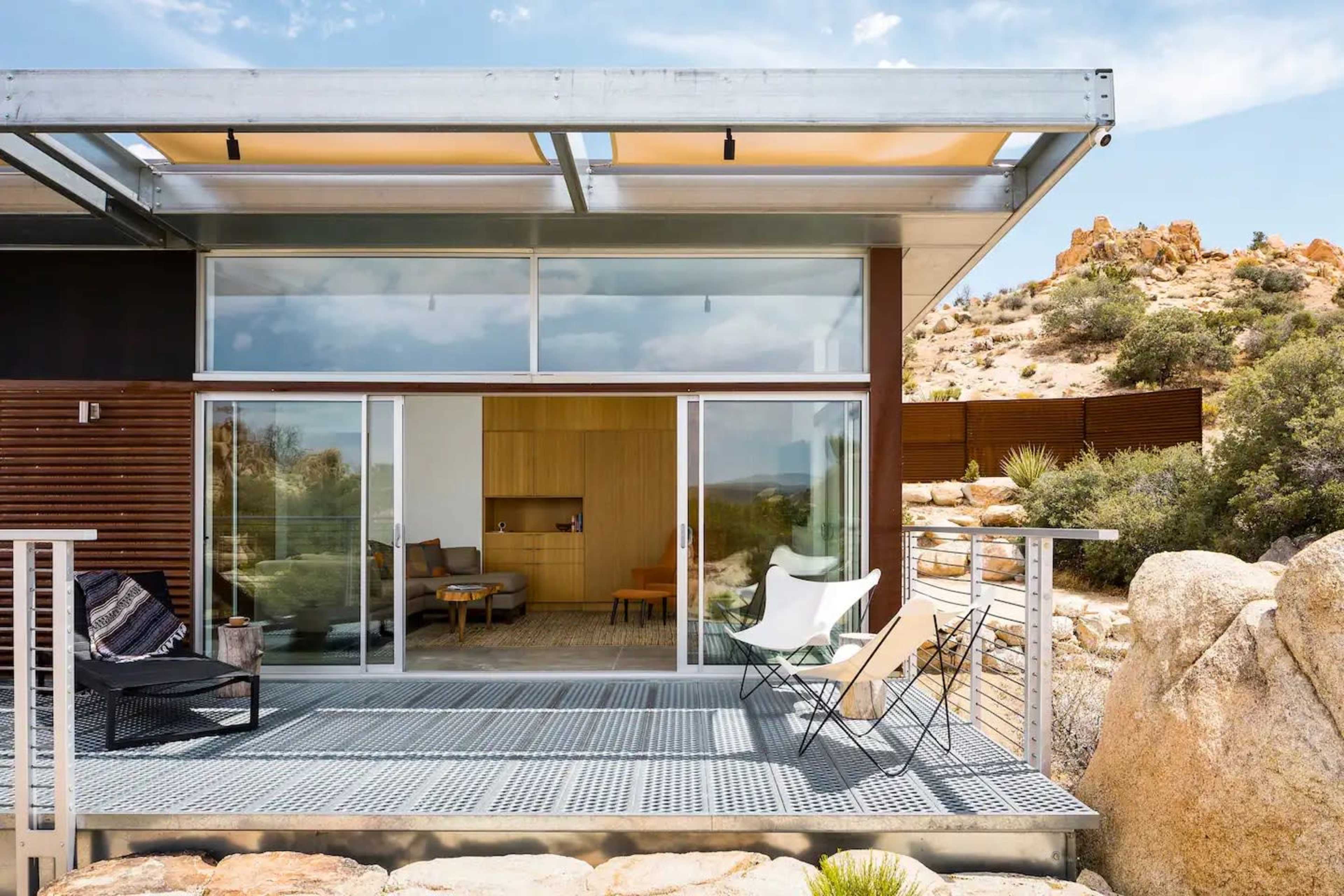 A modern house with large glass doors opens onto a deck surrounded by rocky desert landscape.