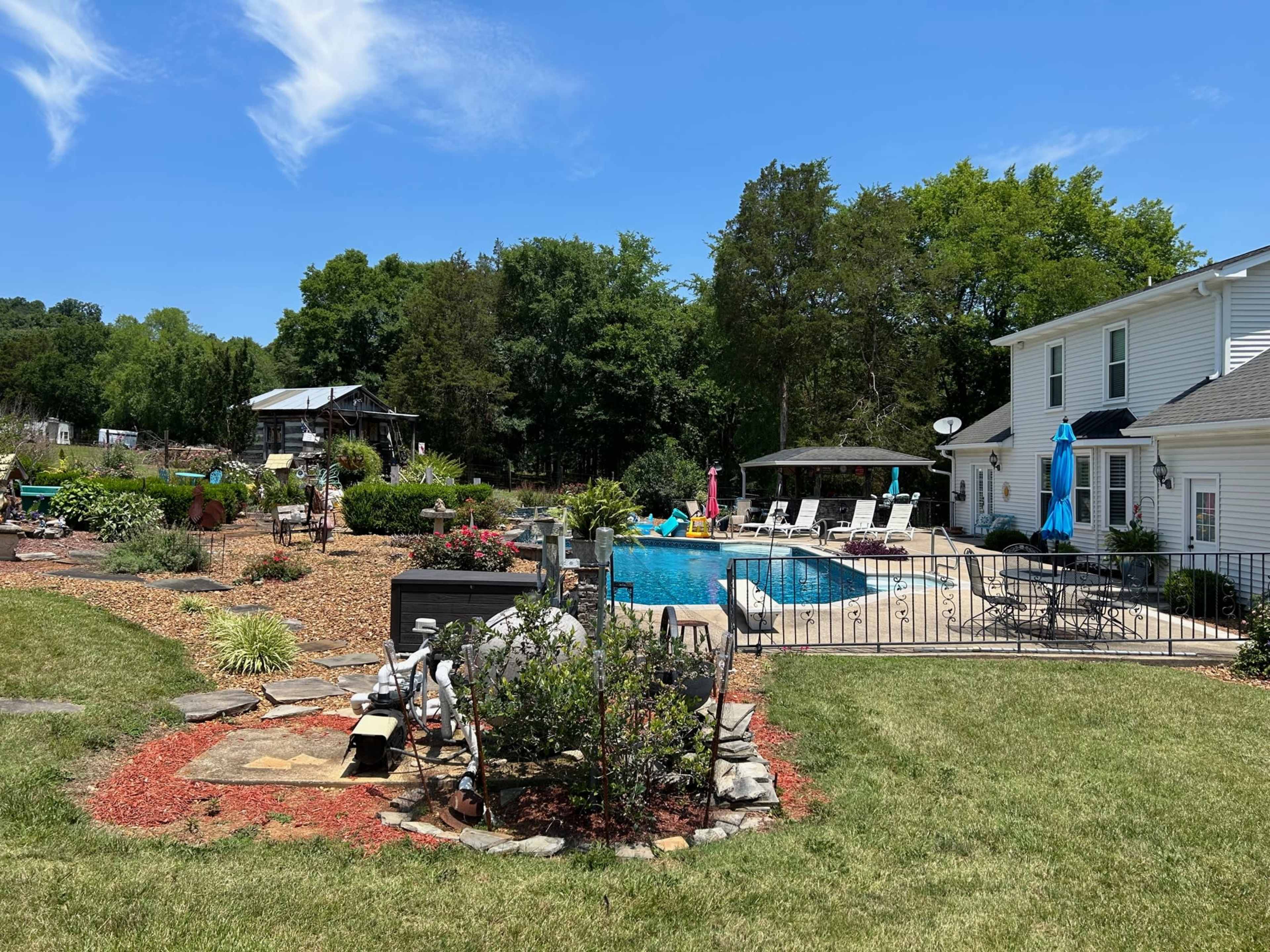 The image shows a landscaped backyard with a swimming pool, surrounded by greenery, garden beds, and outdoor seating areas.