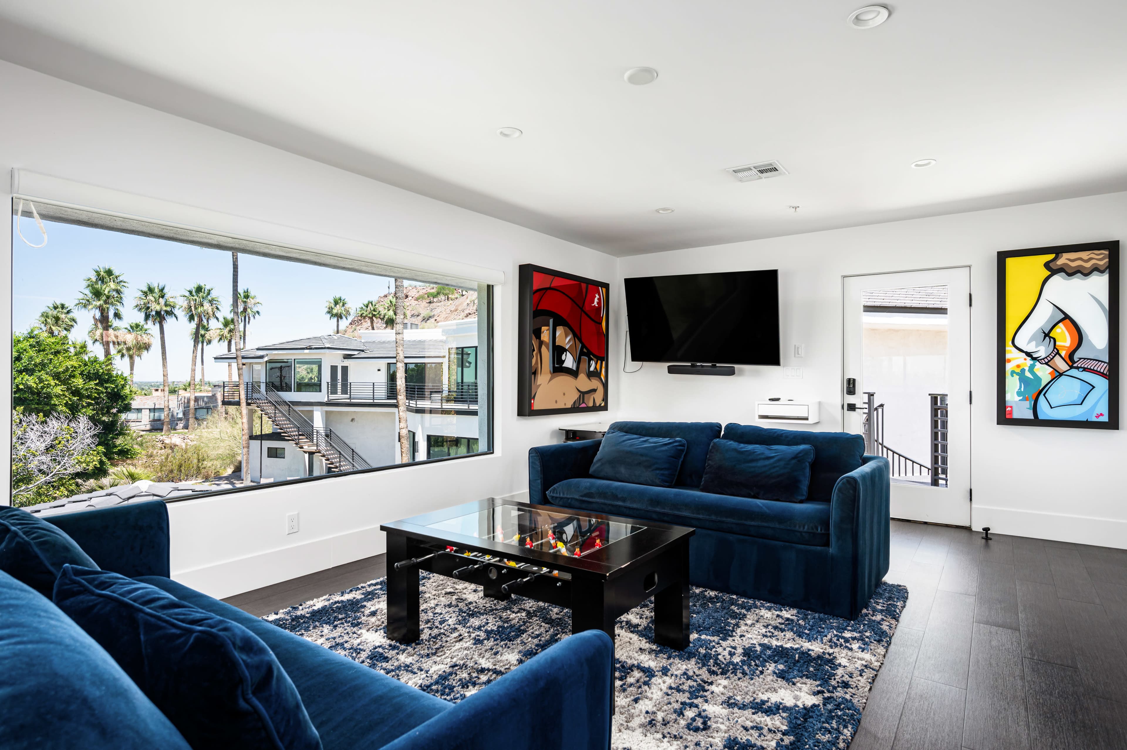 A modern living room features blue sofas and a black coffee table, with large windows providing a view of palm trees and outdoor space.