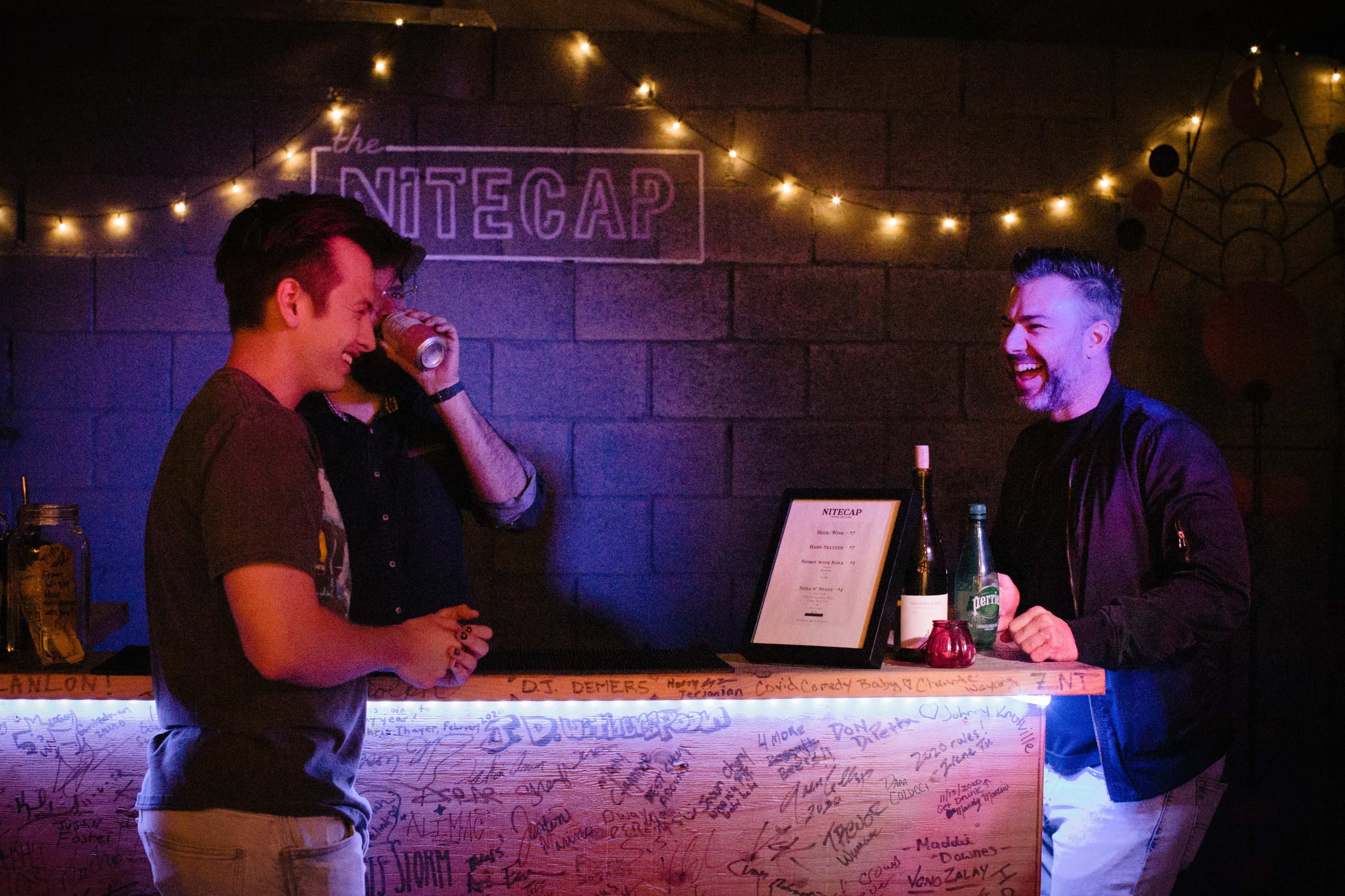 Three men are engaged in conversation at a bar decorated with string lights and a sign that reads "the Nitecap."