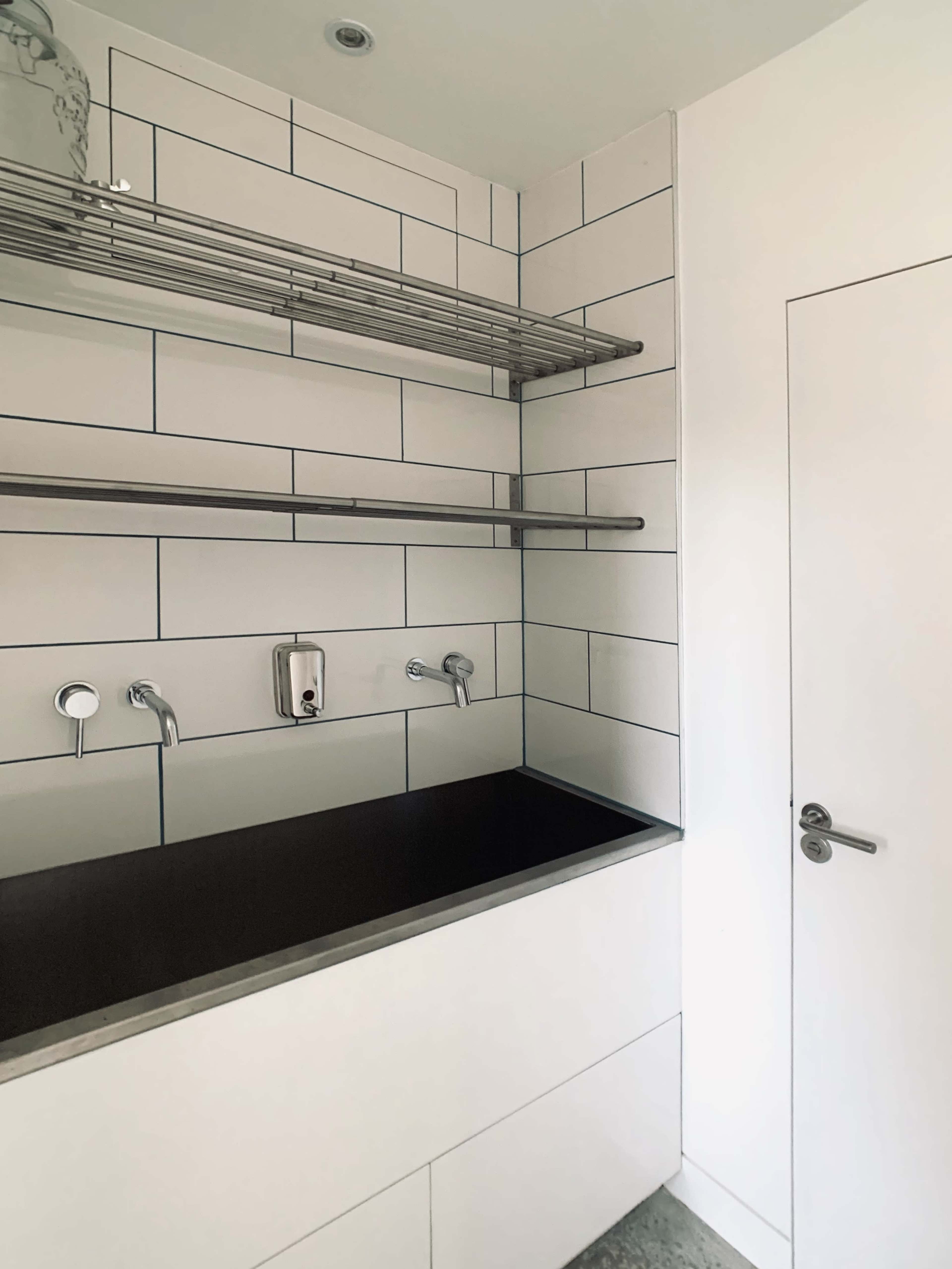 The image shows a clean laundry room with a dark countertop, white tiled walls, stainless steel shelves, and a door on the right.