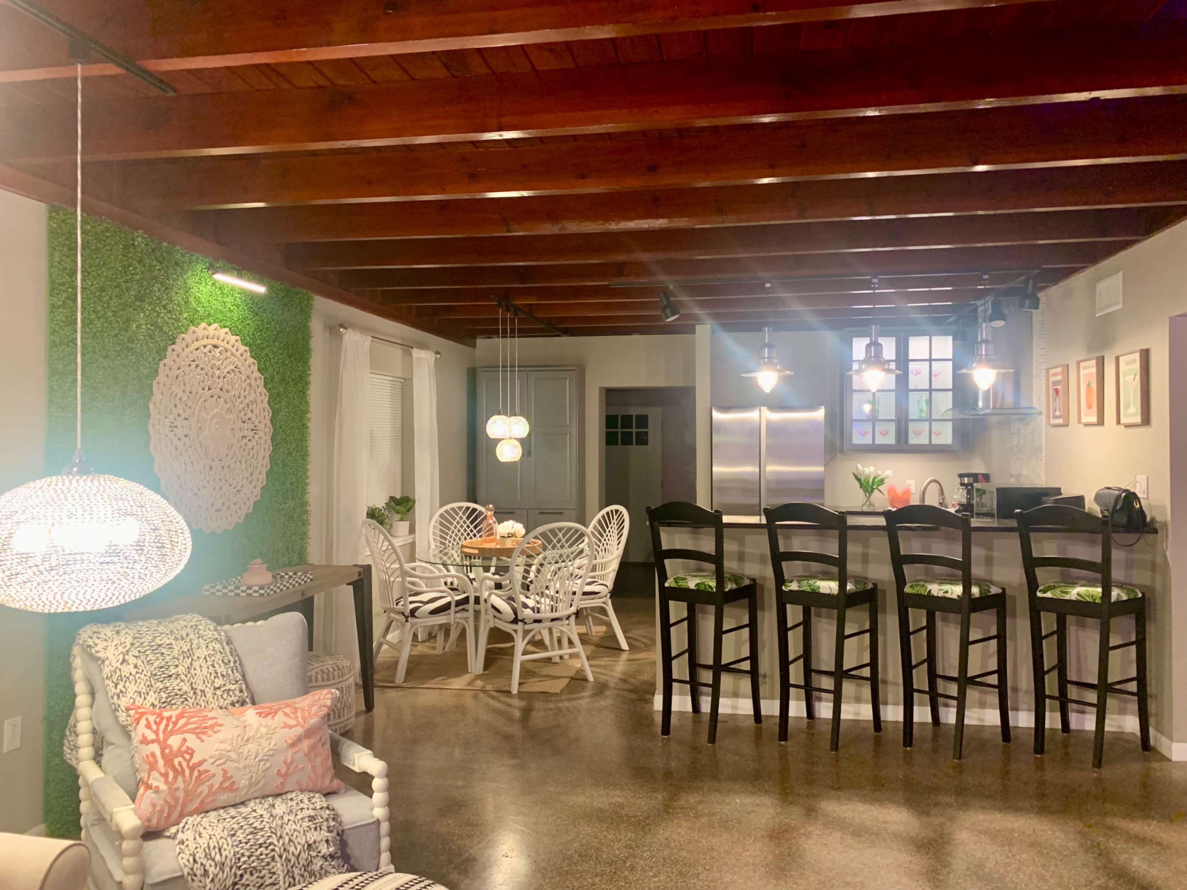 The image shows a modern dining area with wooden beams, a dining table with rattan chairs, and a kitchen visible in the background.