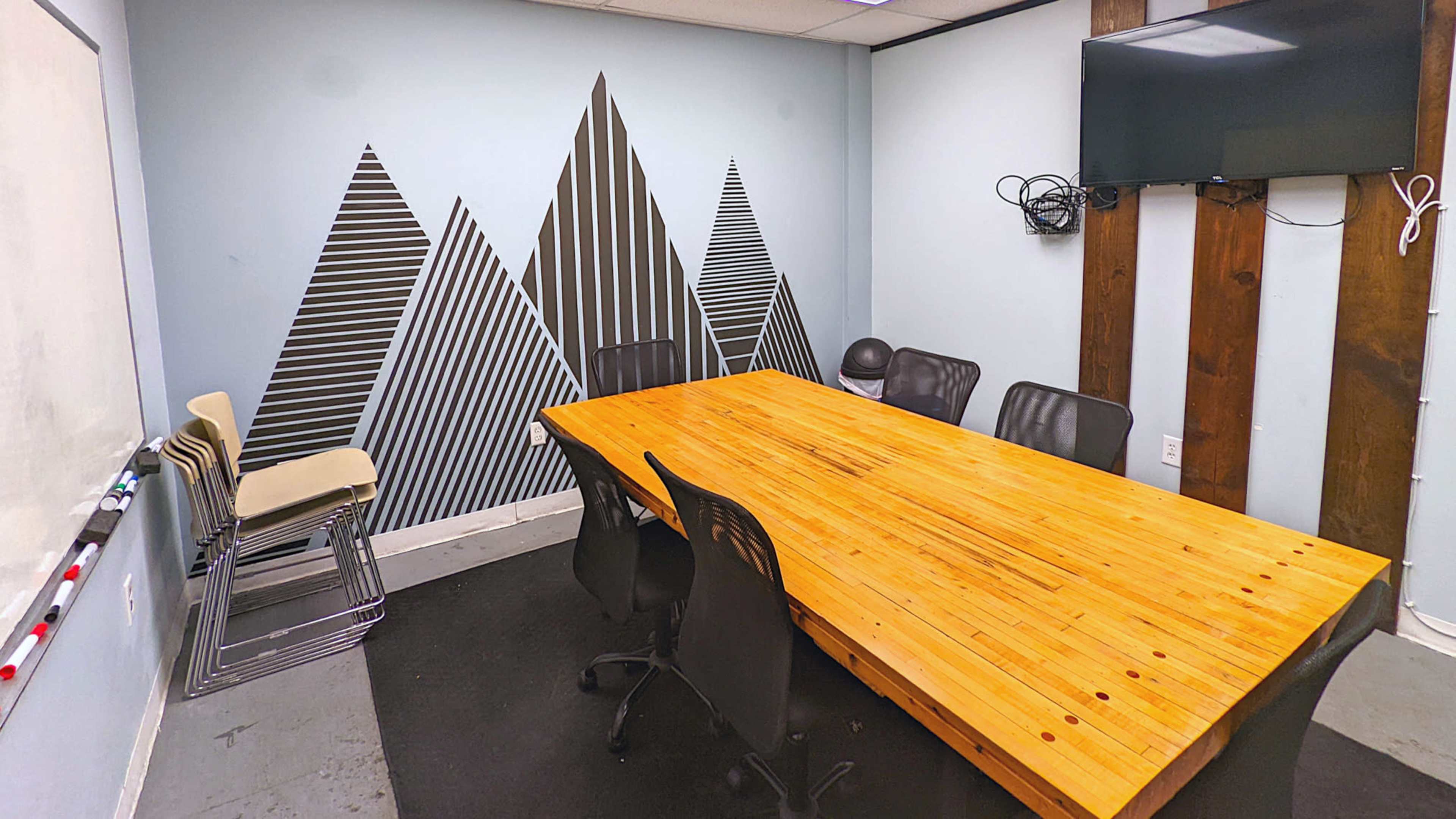 A conference room with a large wooden table surrounded by black chairs, a television mounted on the wall, and a geometric mountain mural on the opposite wall.