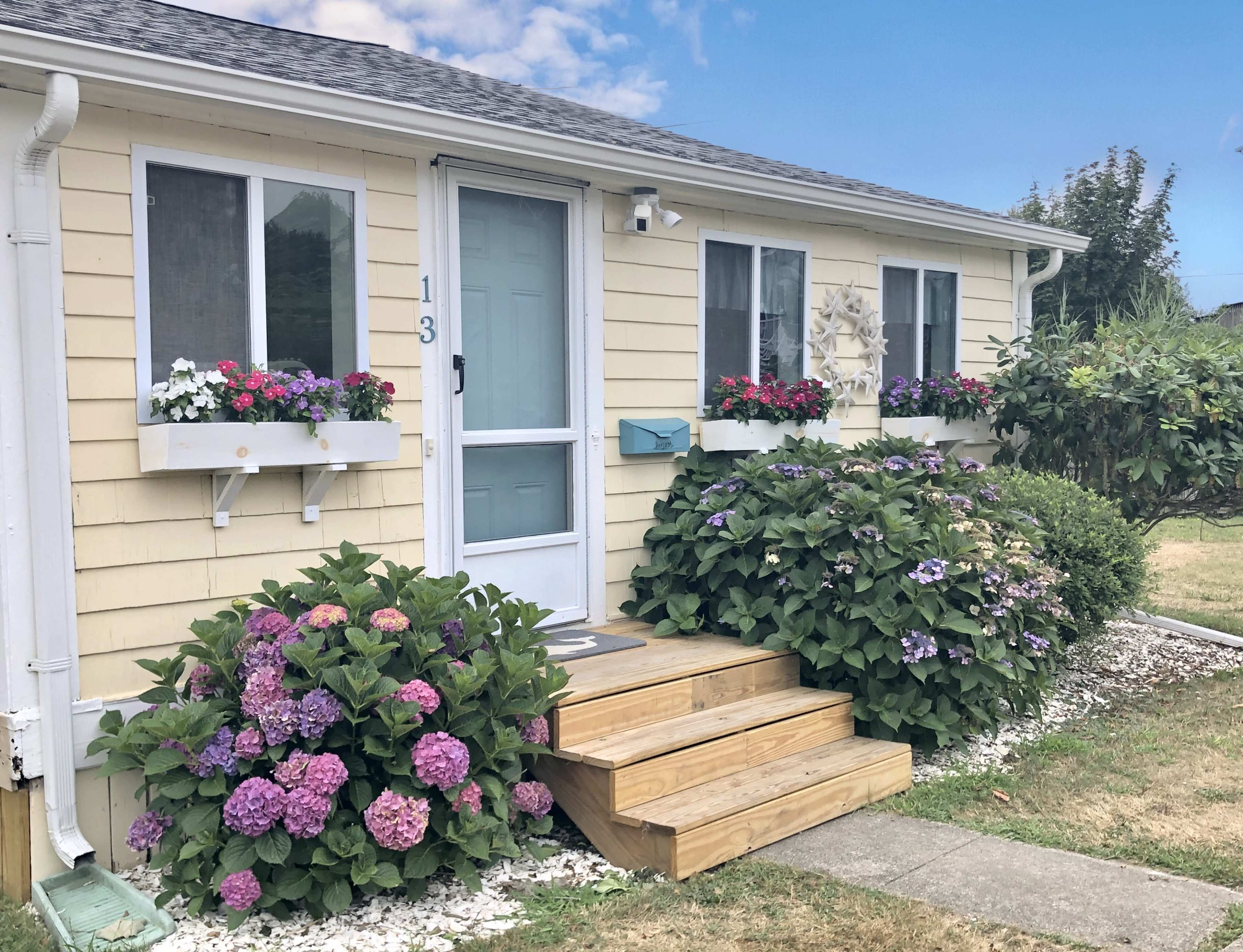 A yellow house with a blue door features wooden steps and planters filled with flowers beside the entrance.