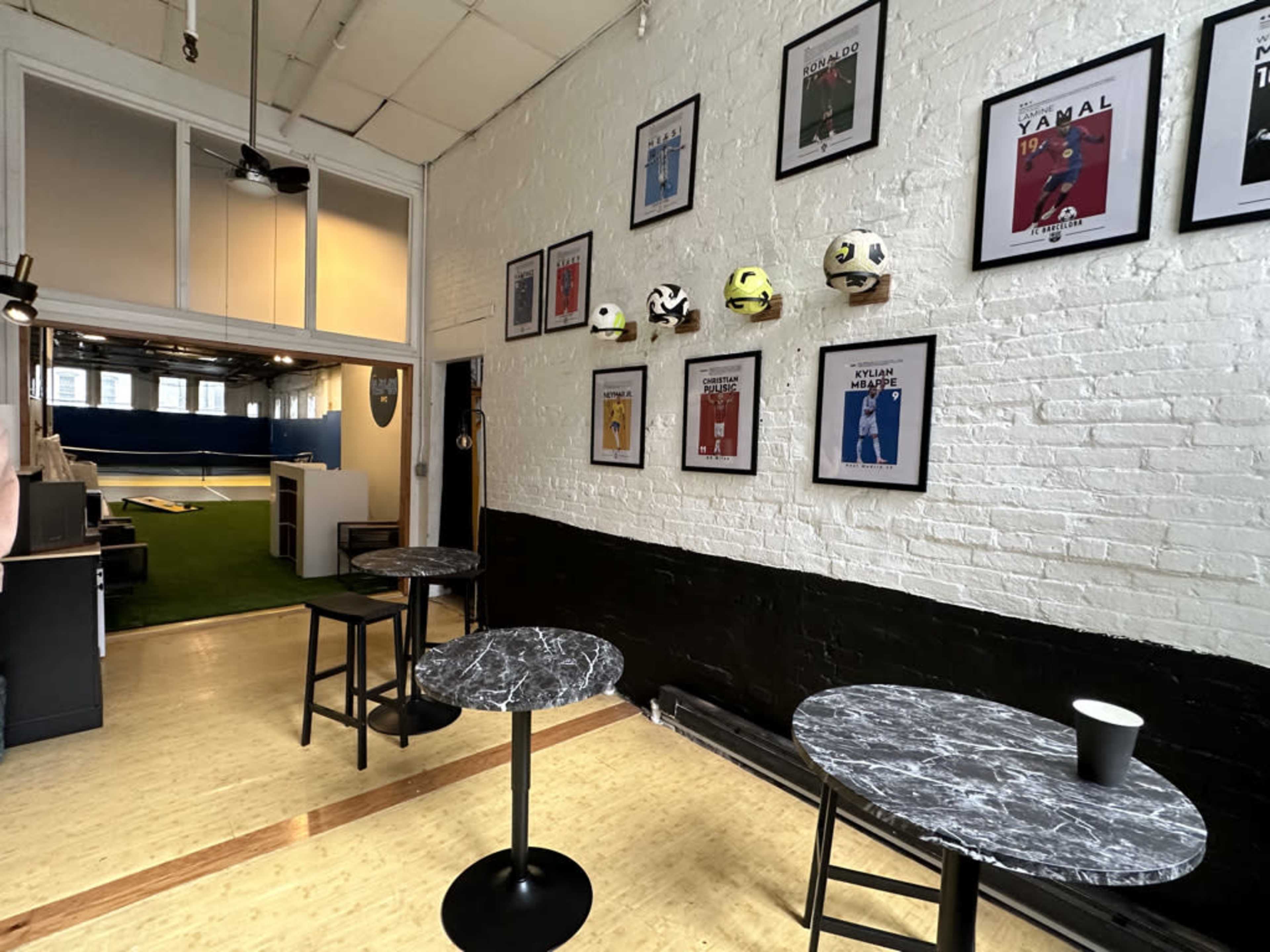 The image shows a room with three marble-topped tables and framed sports posters on the walls, leading to a space with artificial turf and soccer equipment in the background.