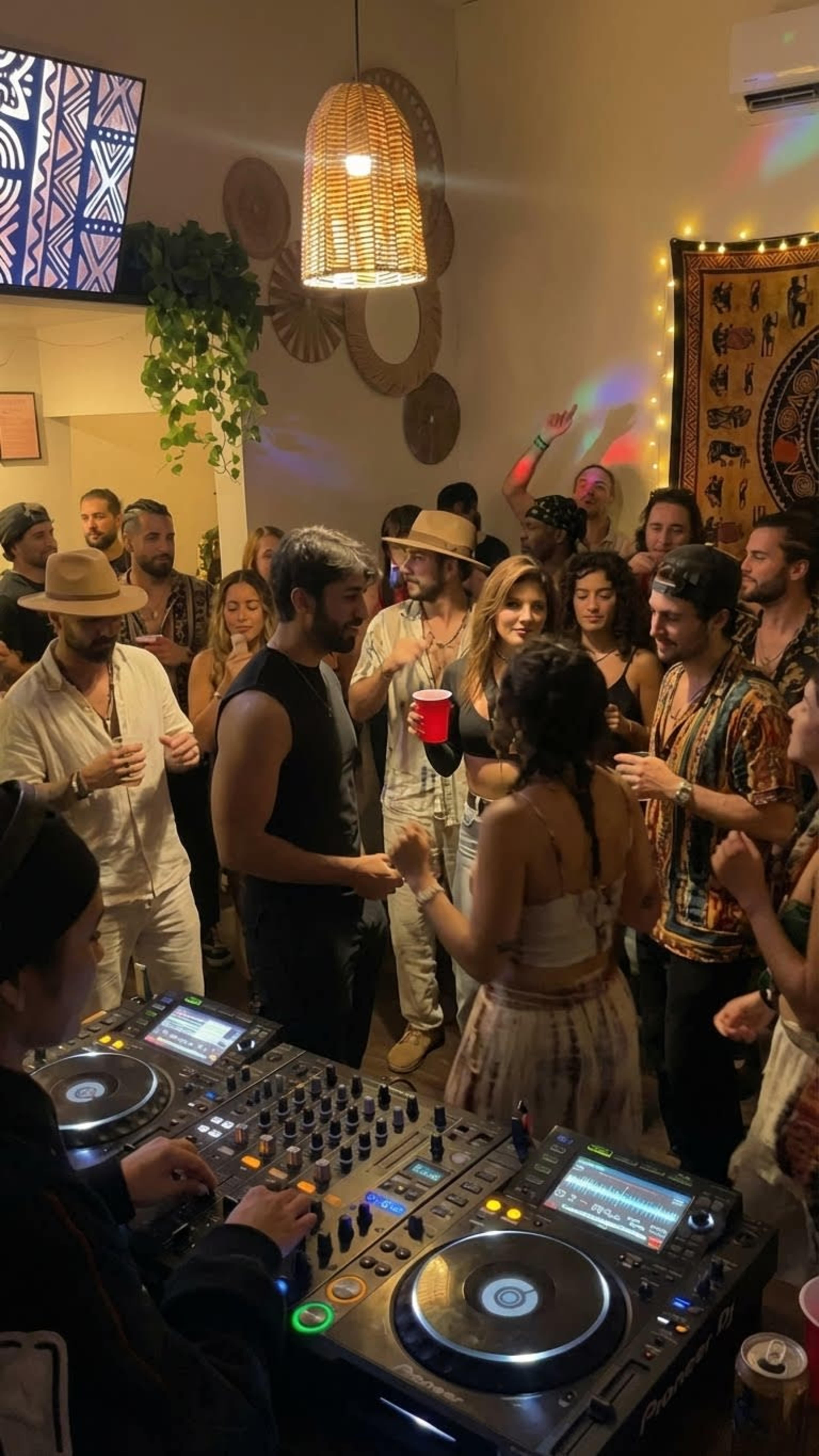 A lively gathering of people is dancing and socializing in a warmly lit room, with two DJs mixing music at the front.