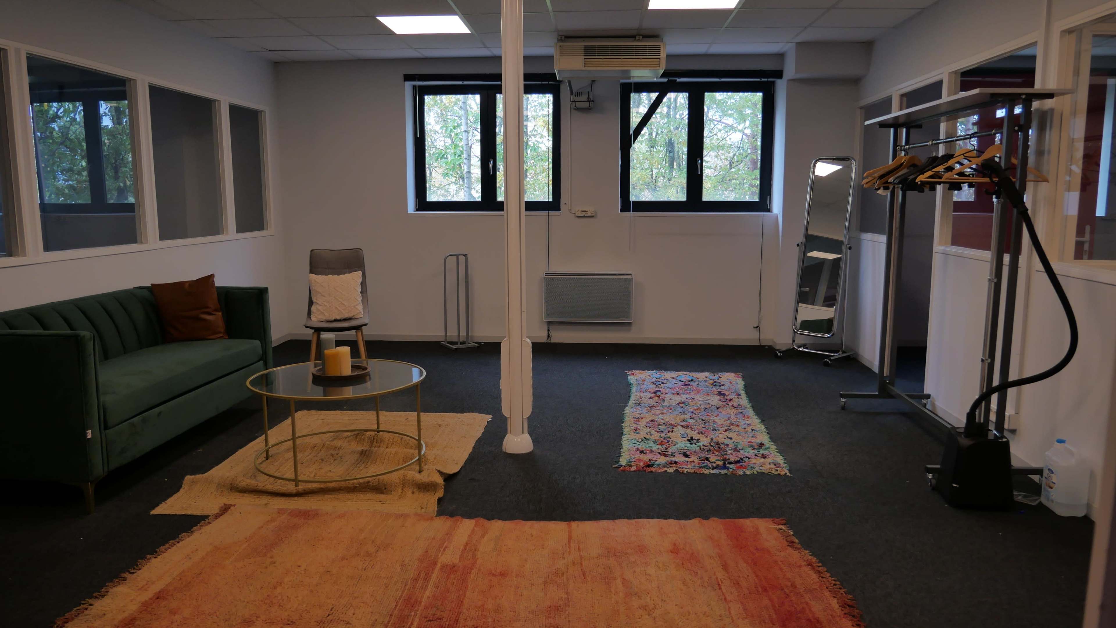 A spacious, well-lit office area featuring a green sofa, a coffee table, a chair, and several rugs on the dark carpeted floor.