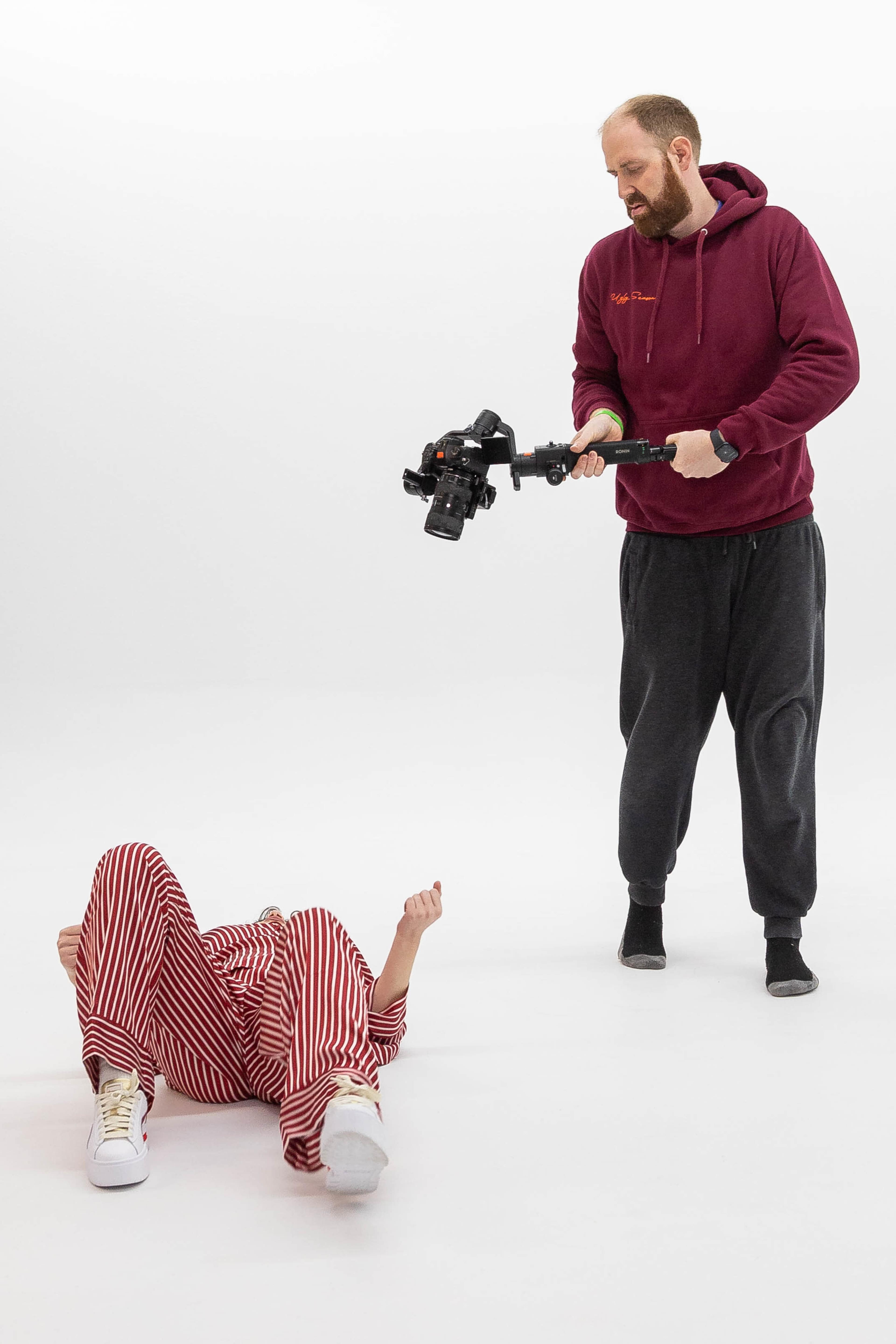A person in a burgundy hoodie holds a camera stabilizer while another individual lies on the ground wearing red and white striped clothing.