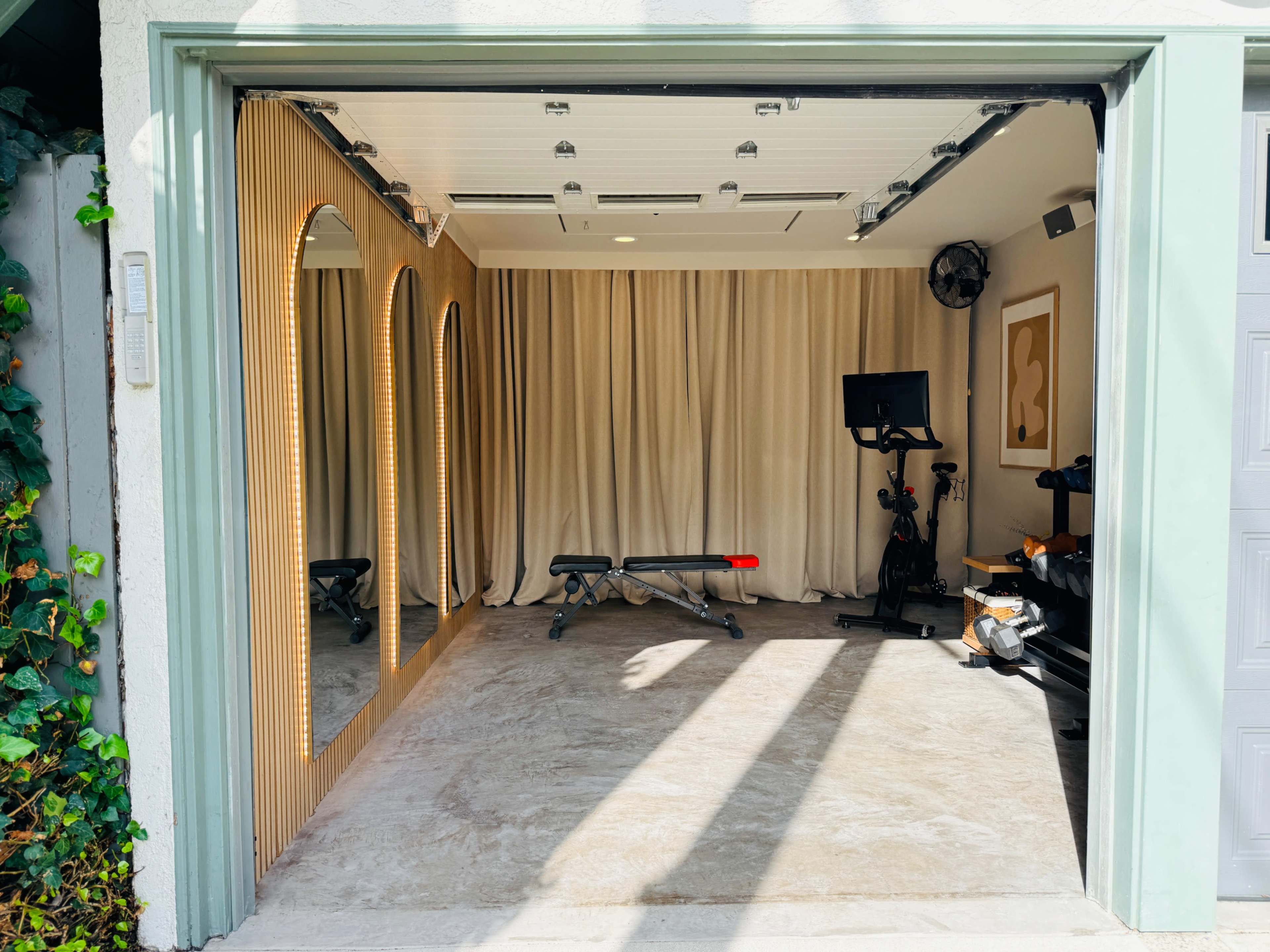 The image shows a garage converted into a home gym, featuring exercise equipment, mirrors framed by wooden arches, and curtains along one wall.