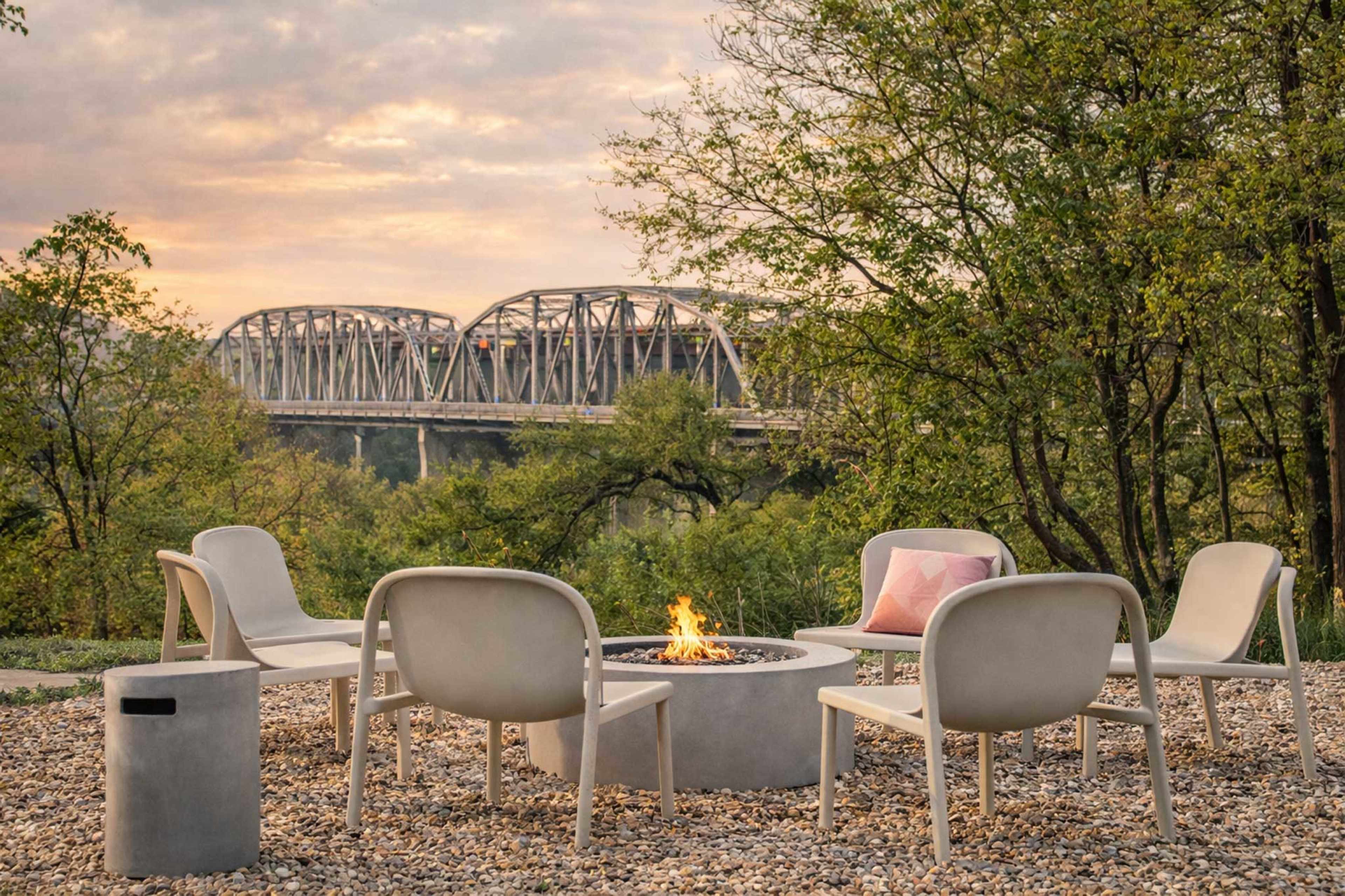 A fire pit surrounded by white chairs sits on a gravel area, with a bridge crossing a river in the background.
