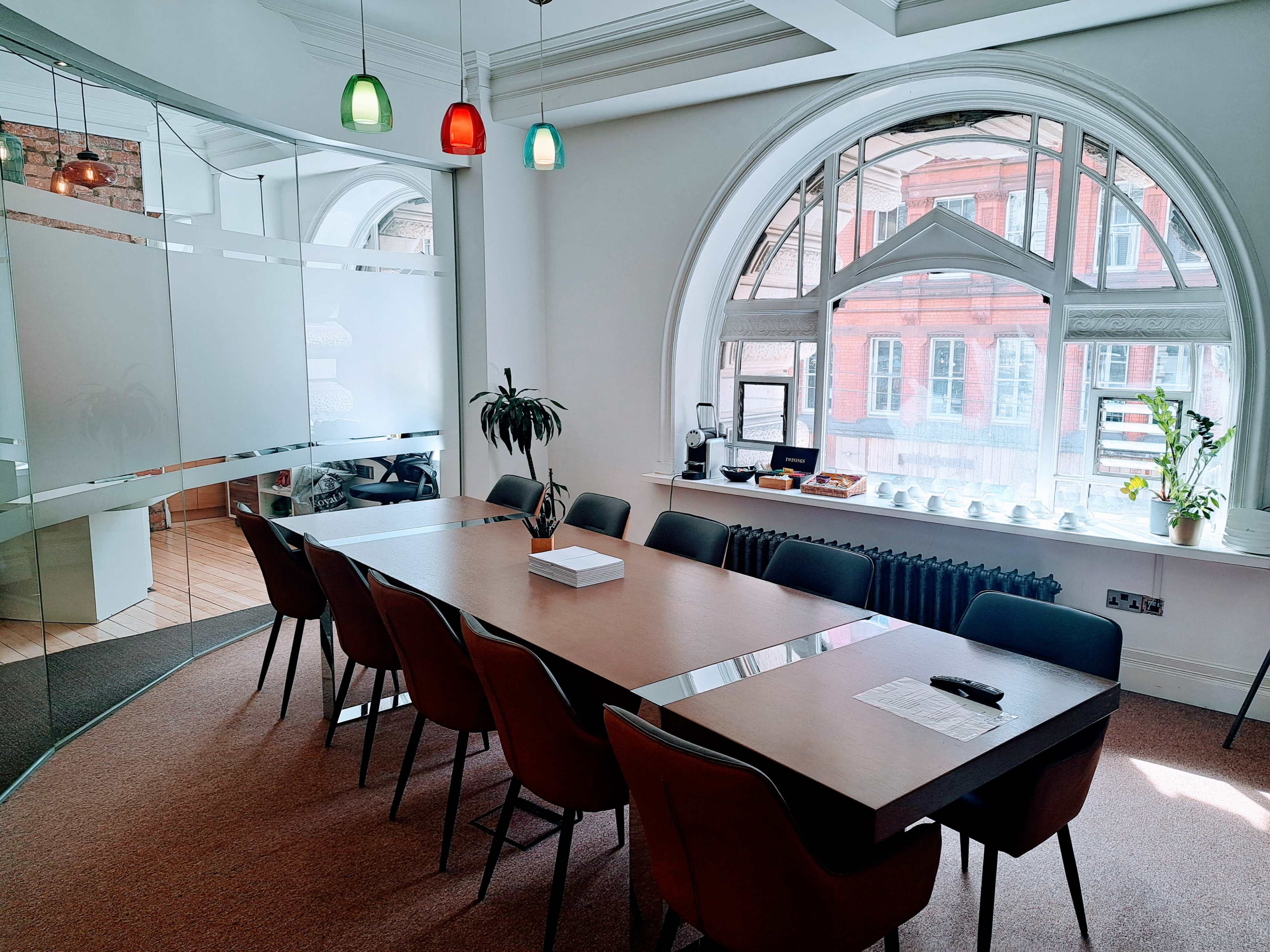 A large conference table surrounded by chairs sits in a bright, modern meeting room with an arched window and colorful pendant lights.