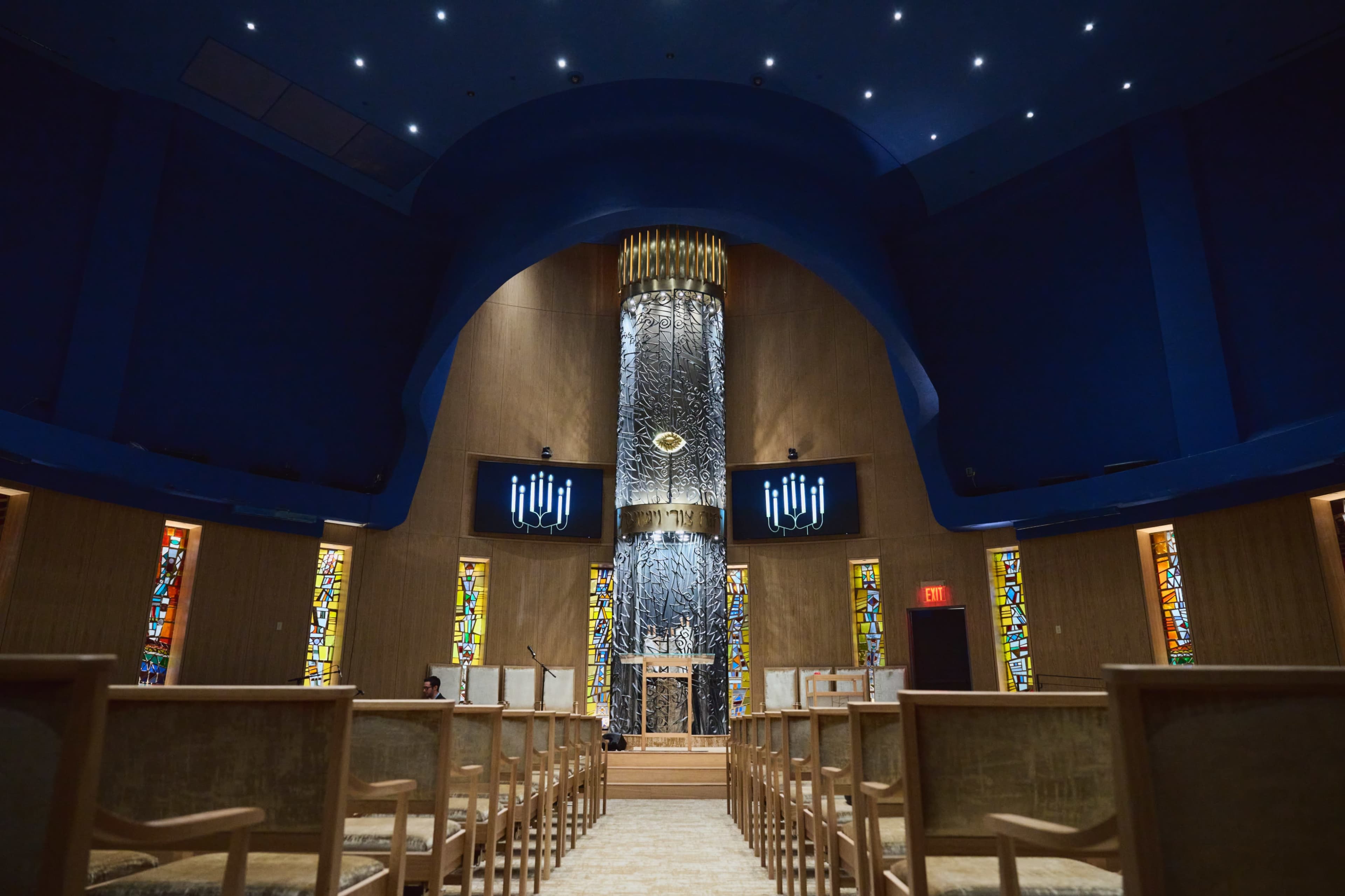 The interior of a synagogue features a central ornate ark surrounded by vibrant stained glass windows and rows of wooden chairs facing forward.