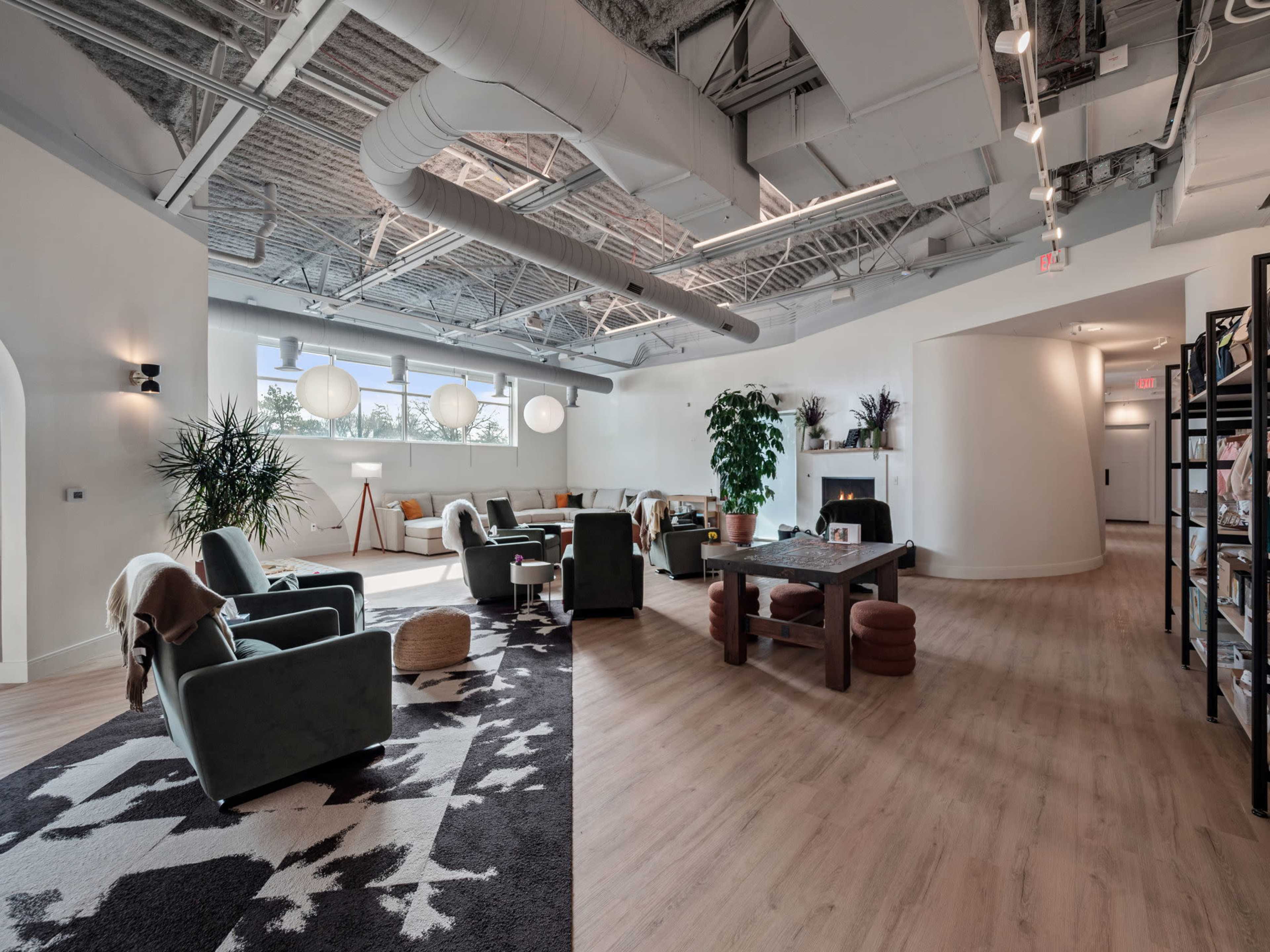 A modern lounge area featuring comfortable seating, large windows, plants, and a communal table.
