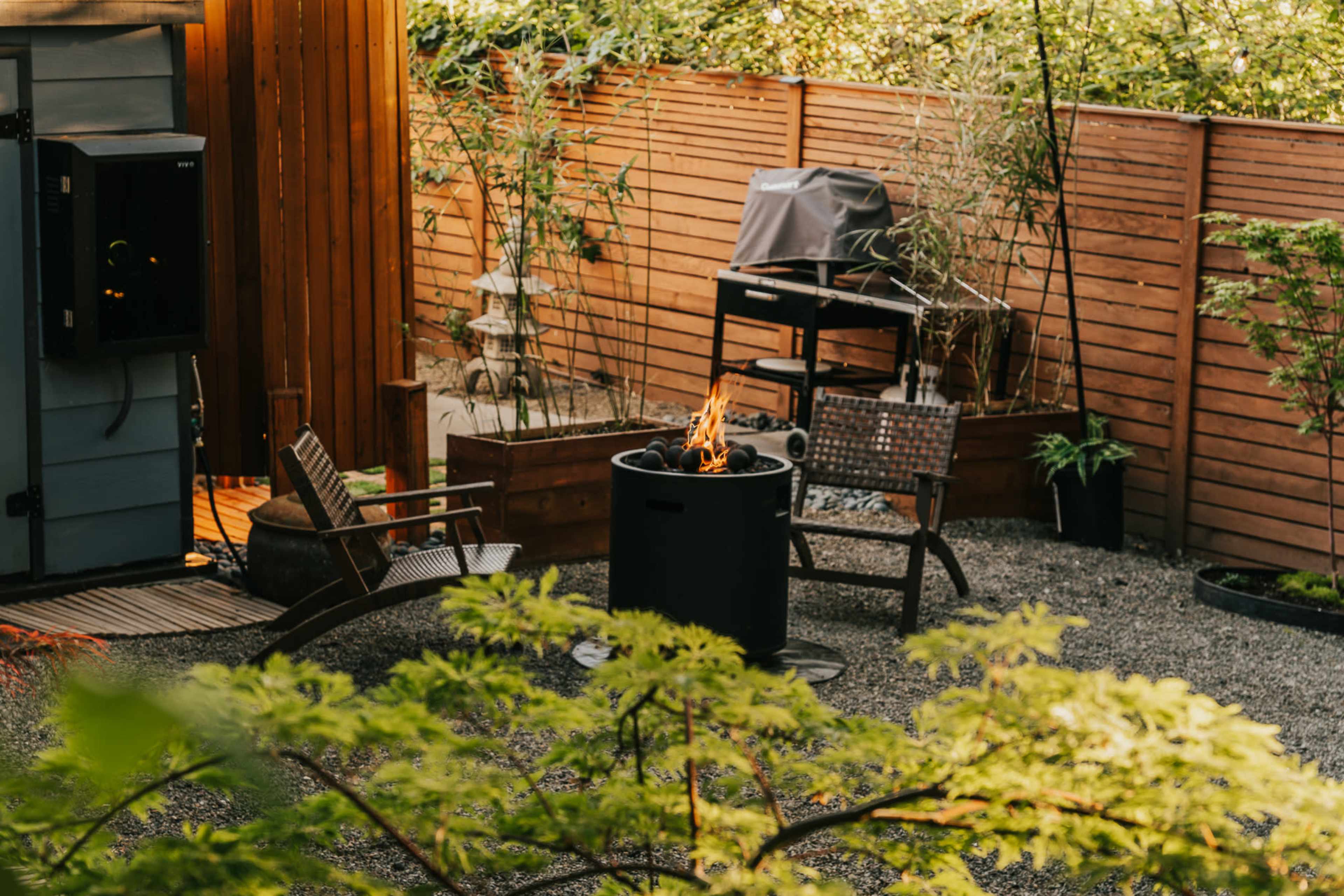 The image shows a cozy outdoor space with a fire pit surrounded by two chairs, gravel flooring, and a grill in the background, framed by bamboo and wooden fencing.