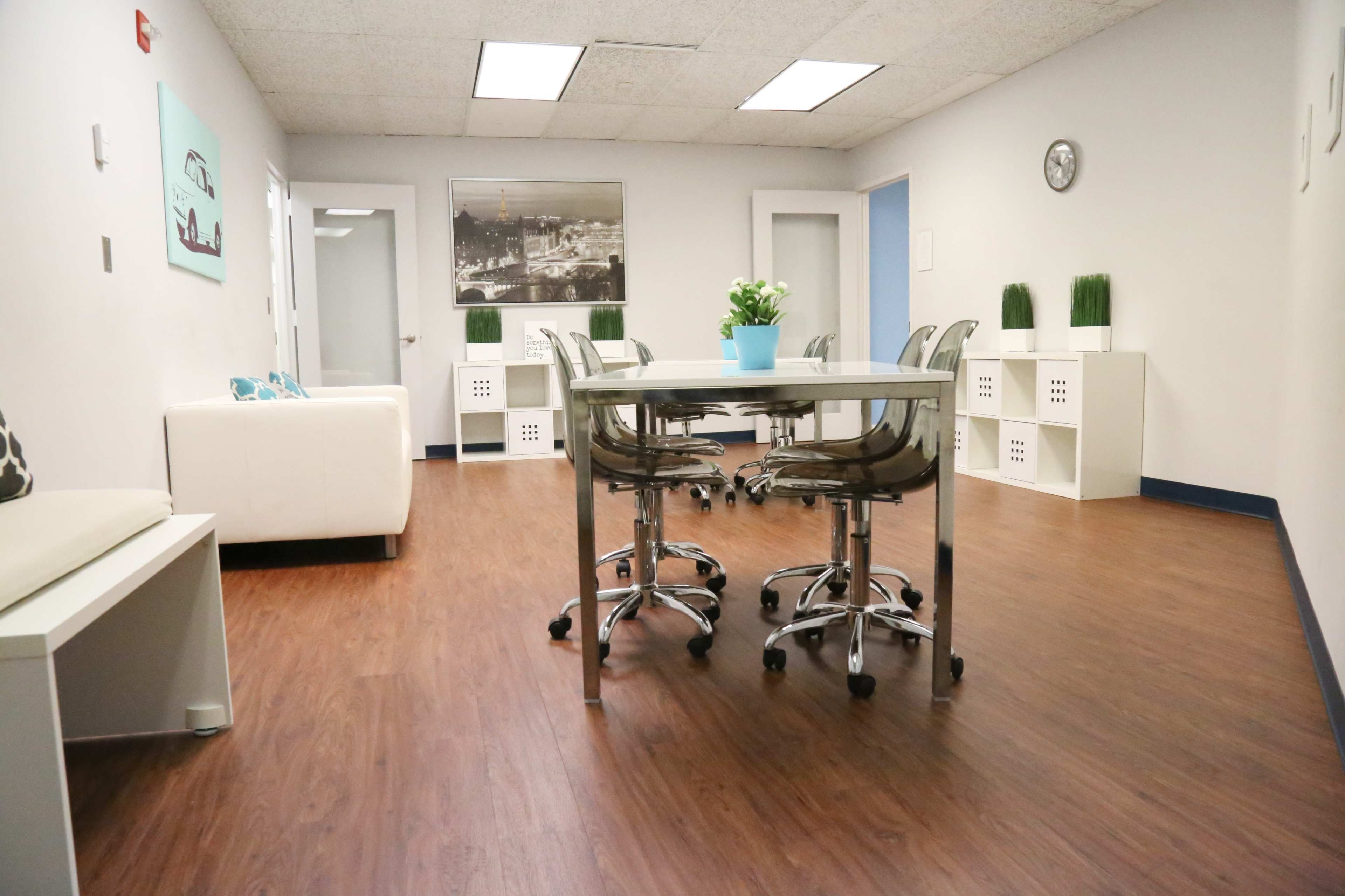 A modern office space features a white table surrounded by clear chairs, with a potted plant on the table and light-colored walls.