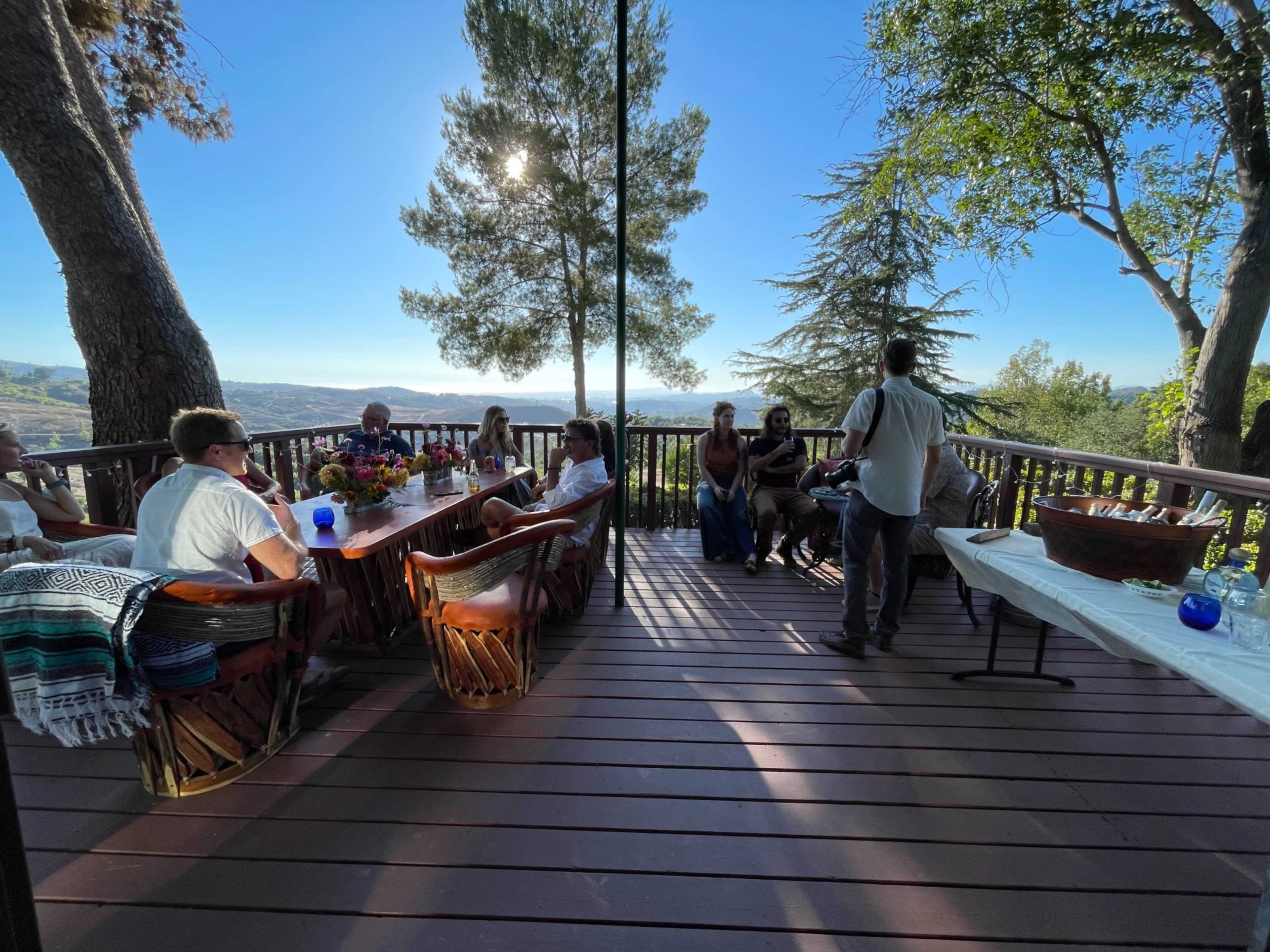 A group of people is seated at a table on a wooden deck overlooking a scenic landscape.