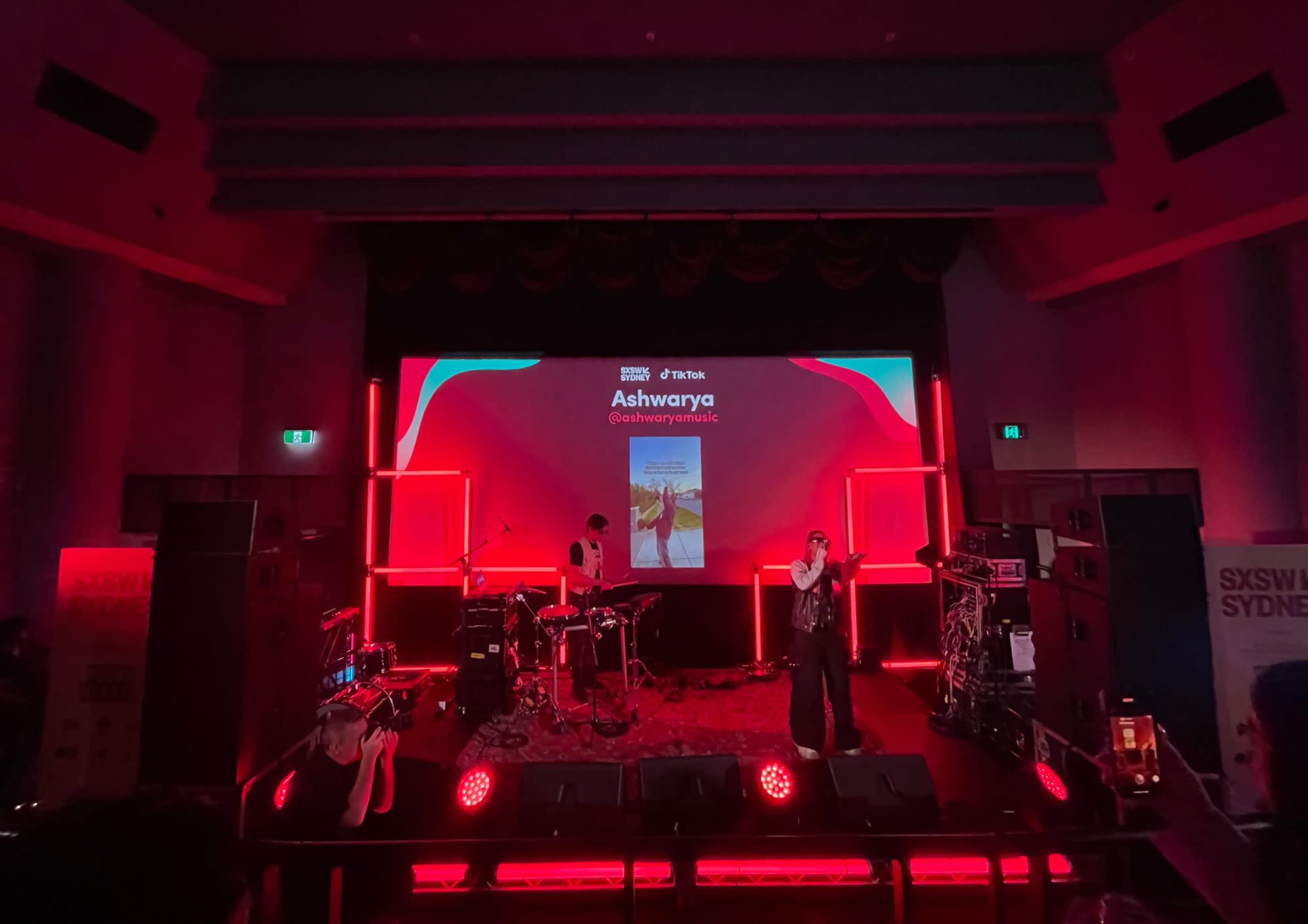 A performer stands on stage in front of a large screen displaying their name and social media handle, with instruments set up for a band in the background and red lighting illuminating the scene.