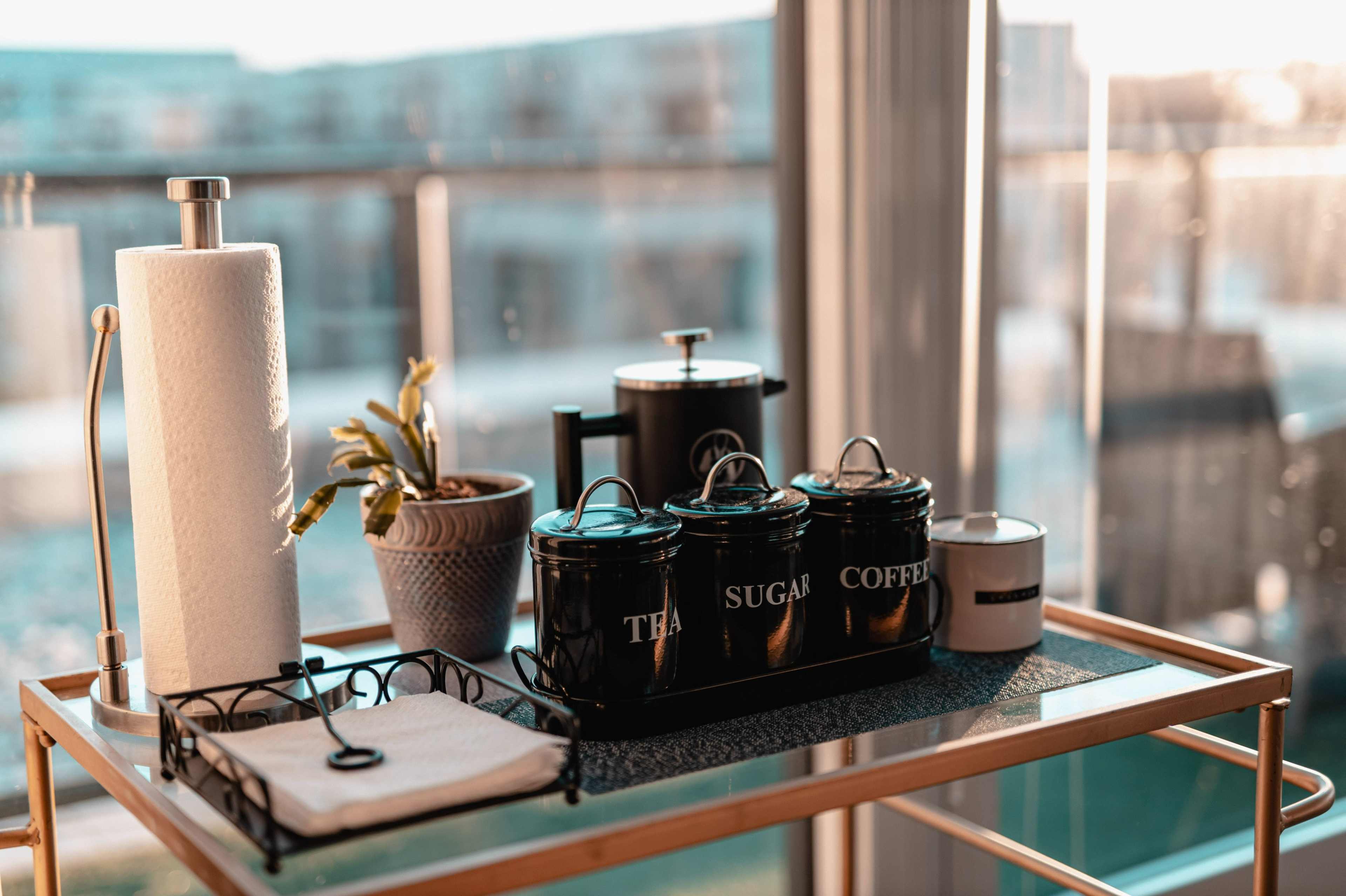 A golden cart displays various kitchen items, including three black canisters labeled "Tea," "Sugar," and "Coffee," a potted plant, a coffee maker, and a candle, all set against a backdrop of large glass windows.