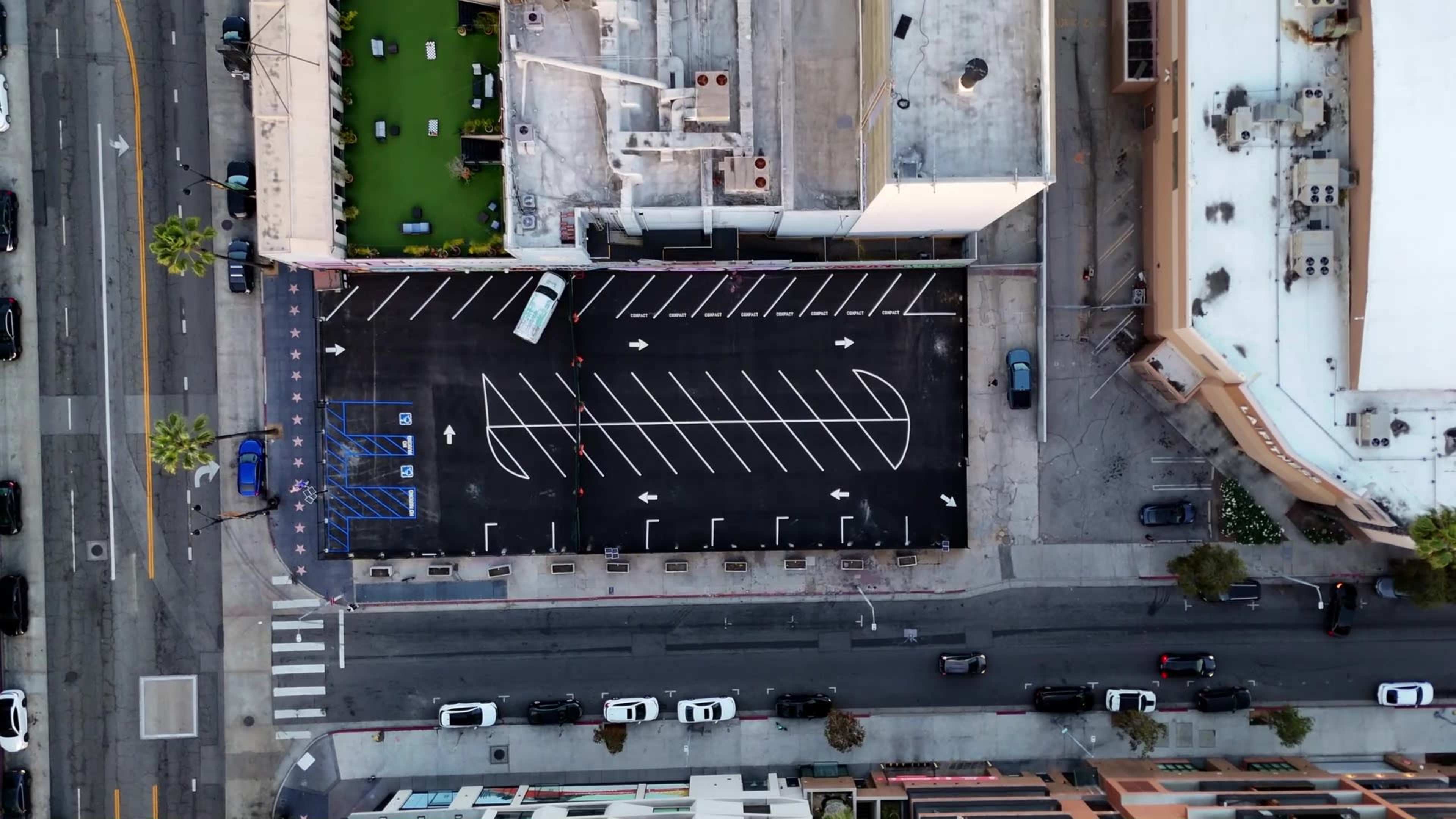 The image shows an aerial view of a parking lot with marked spaces alongside a street lined with vehicles and buildings.