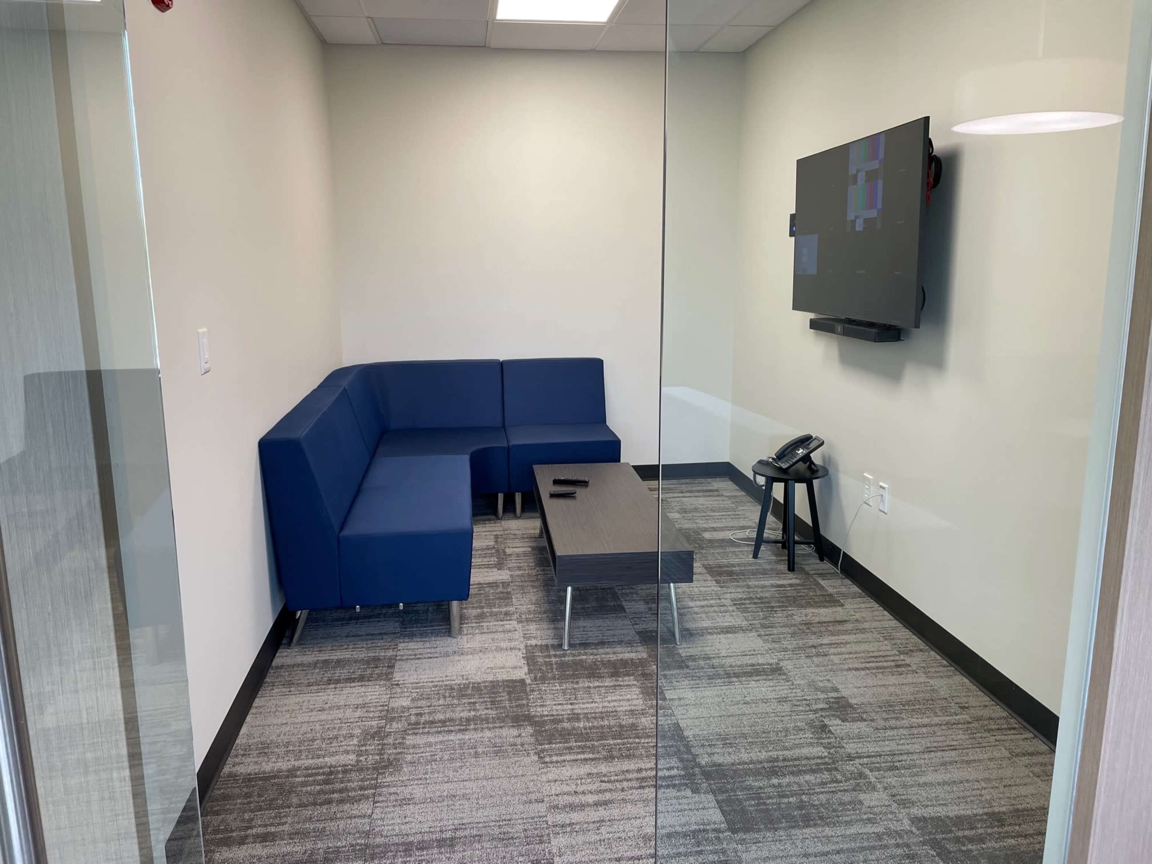 A small, modern meeting room featuring a blue sectional couch, a black coffee table, a wall-mounted television, and a telephone.